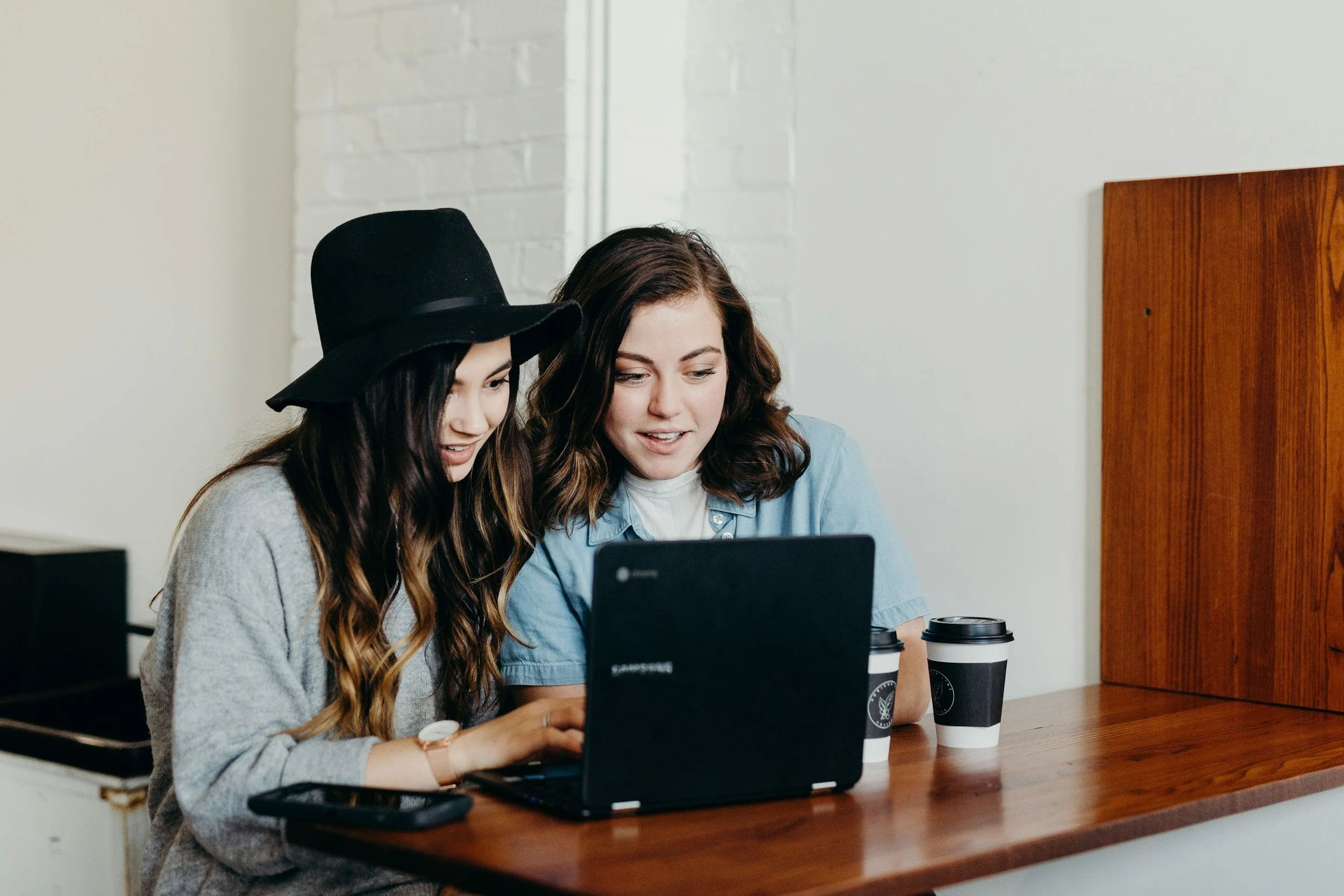 Two women sitting at a wooden table with coffee cups, looking at a laptop together.