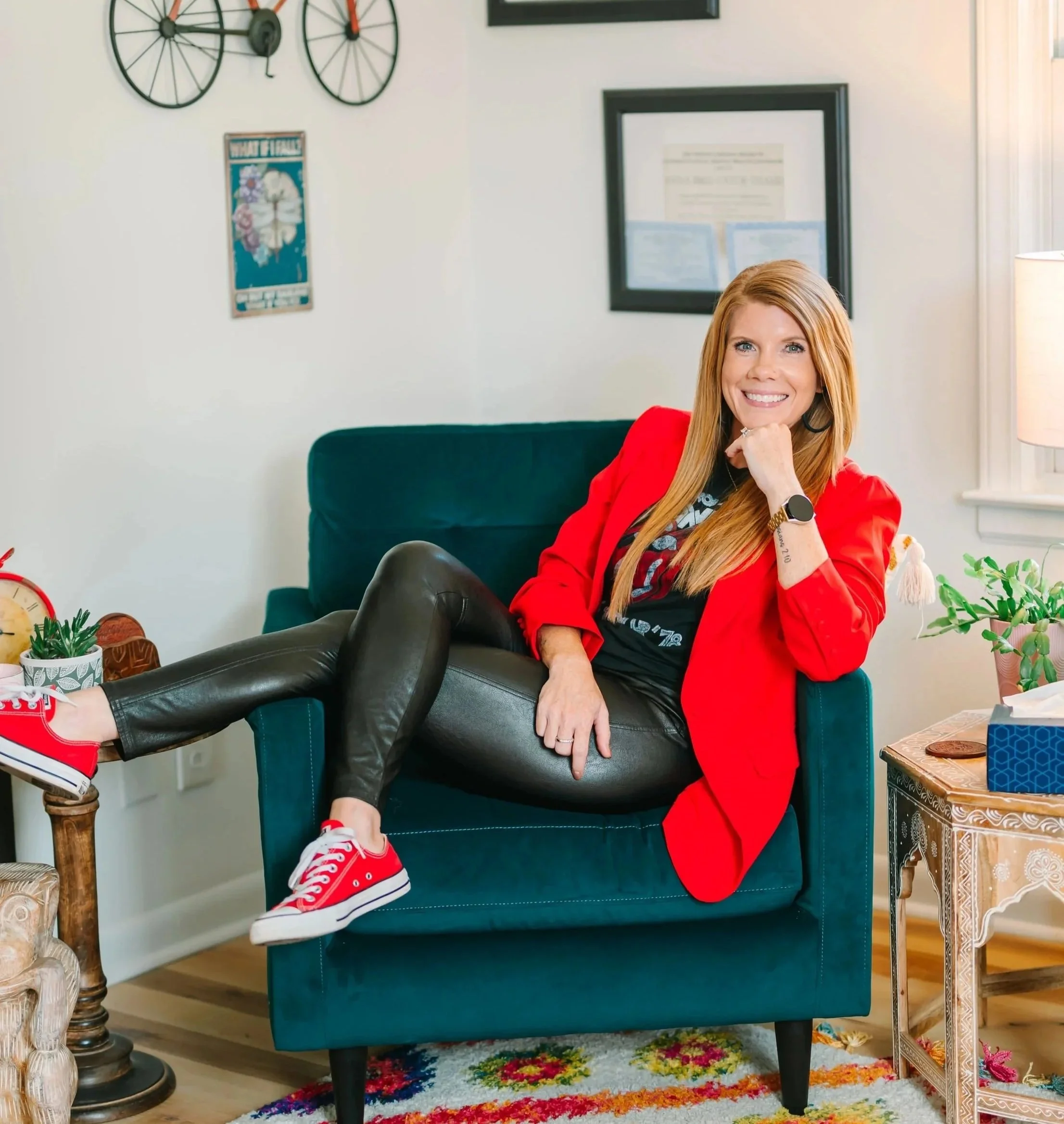 A woman with long red hair smiling and sitting on a teal armchair in a cozy living room. She's wearing a red blazer, black pants, red sneakers, and has a smartwatch on her left wrist. The room has a colorful floral rug, potted plants, framed pictures, a vintage clock, and decorative items on a side table.