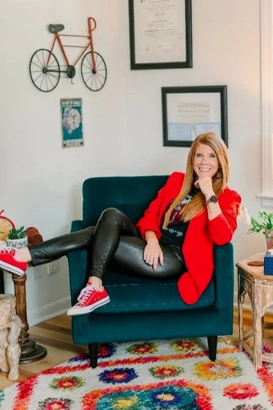 A woman with long blonde hair, wearing a red blazer, black leather pants, and red sneakers, sitting on a teal armchair in a living room, smiling with her leg crossed over the armrest.