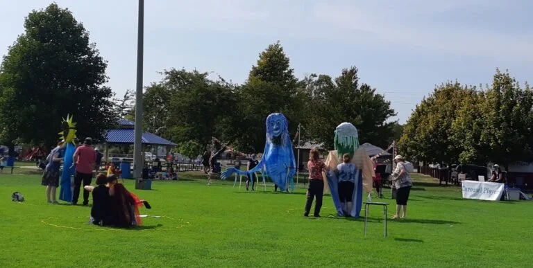 An outdoor scene, three larger than life puppets (puppeteer by at least two people) are in a filed. Audience is dispersed between the puppets