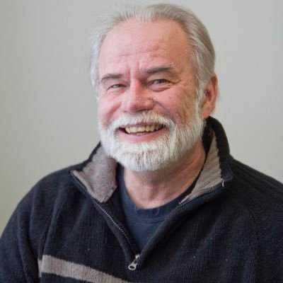 Patrick smiles broadly at the camera with a happiness that is apparent. He is wearing a dark grey top with the collar open.