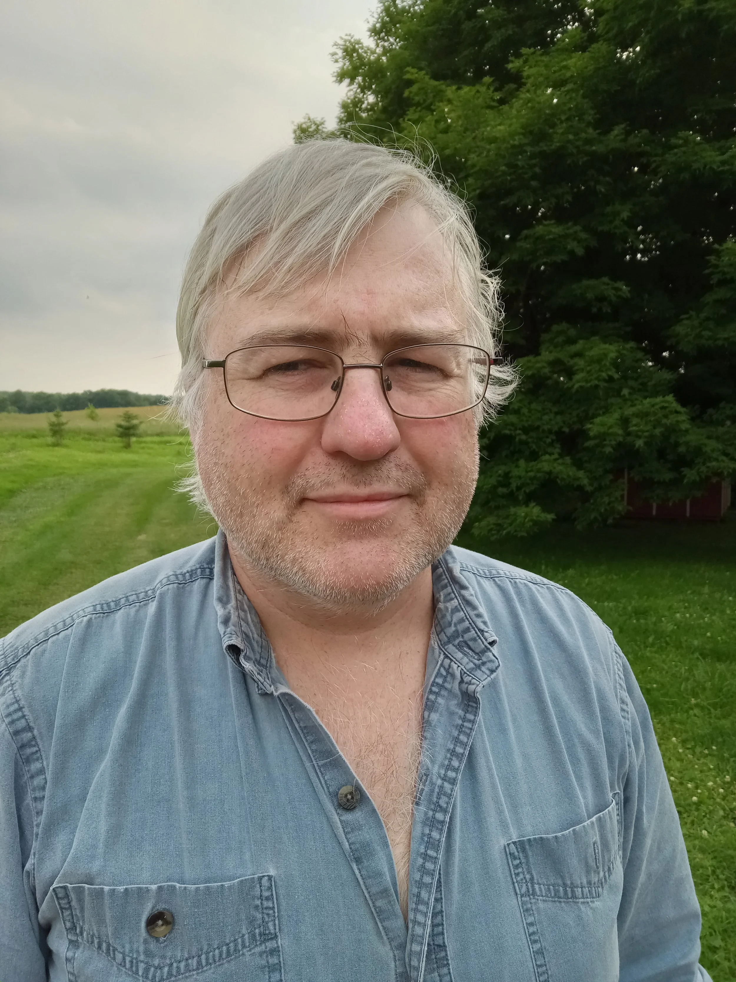Eric stands in a field and looks at the camera with a small smile on his face. He wears glasses and a blue chambray shirt.