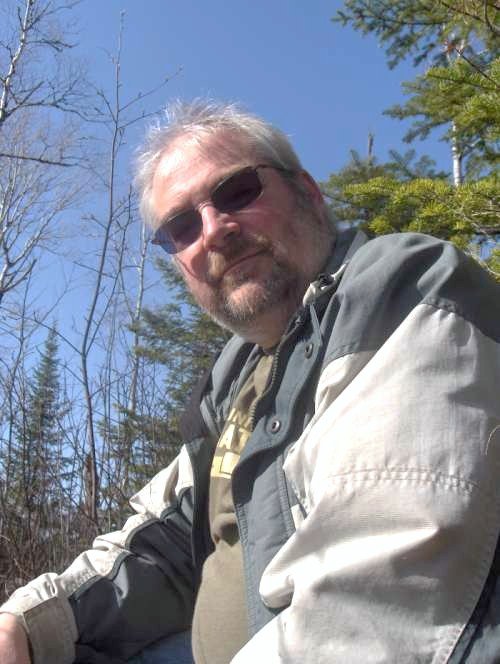 CB is outside in nature. He wears sunglasses and looks down, smiling, at the camera.