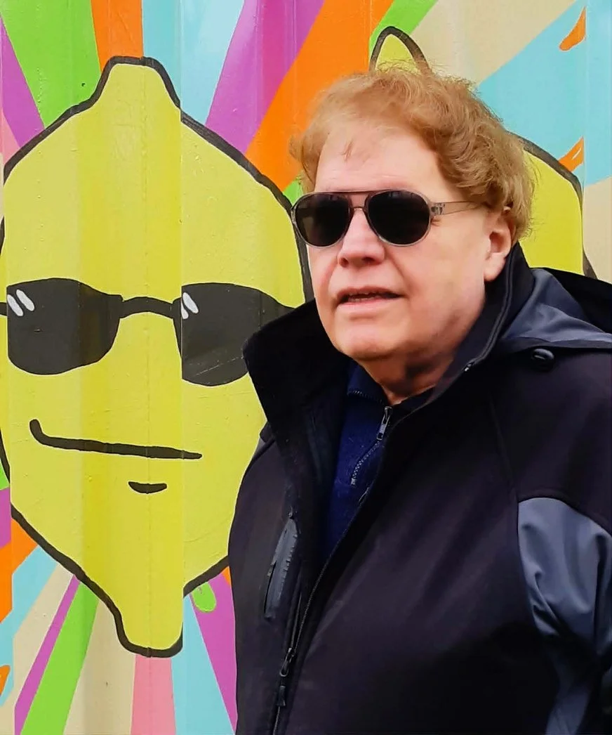 Richard stands in front of a brightly coloured background. He wears sunglasses and looks at the camera in profile.