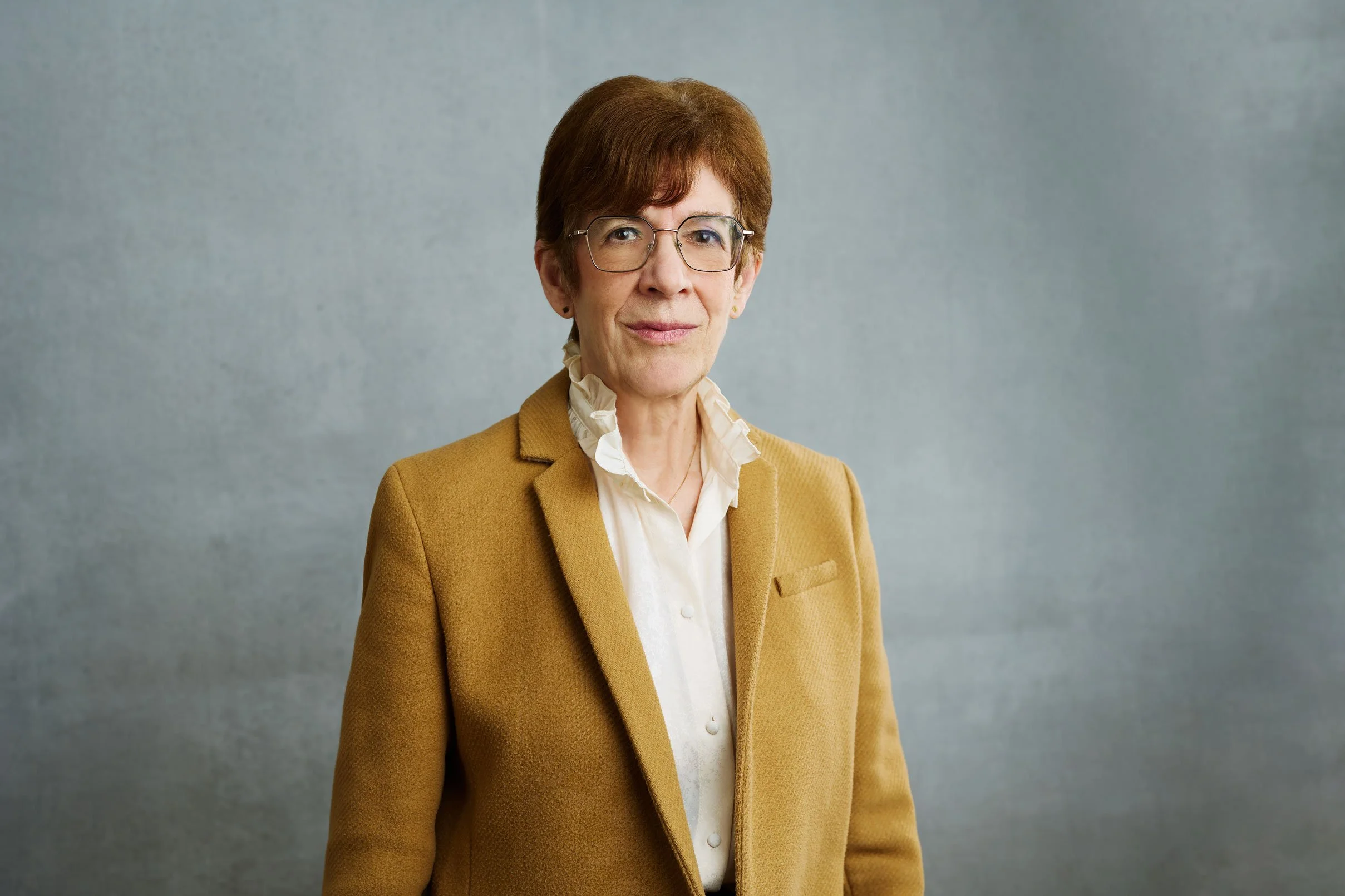 Diane faces the camera. Her short, red hair is combed back and she wears glasses.Shhe is wearing a mustard yellow blazer.