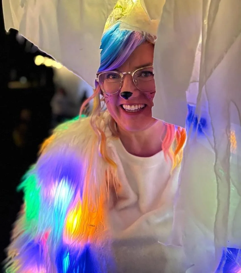 L'artiste, qui sourit à travers plusieurs couches de tissu blanc translucide, porte des lunettes, du maquillage animalier discret et une perruque pastel aux couleurs de l'arc-en-ciel.