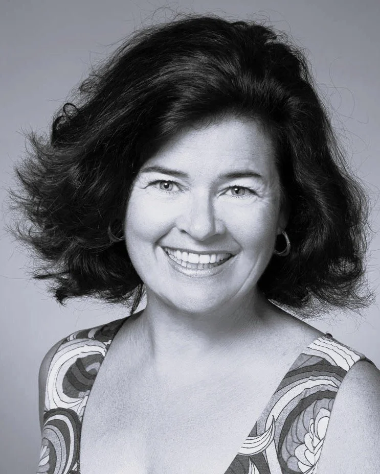 A black and white photo of Colleen. She smiles broadly into the camera. Her dark hair is swept to one side.