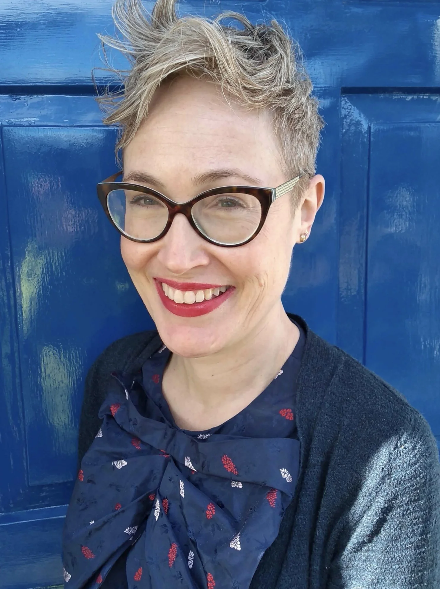 Headshot of Lisa Poushinsky, a smiling white middle-aged woman with glasses and short light hair, wearing a navy blue blouse and sweater, standing in front of a blue door.