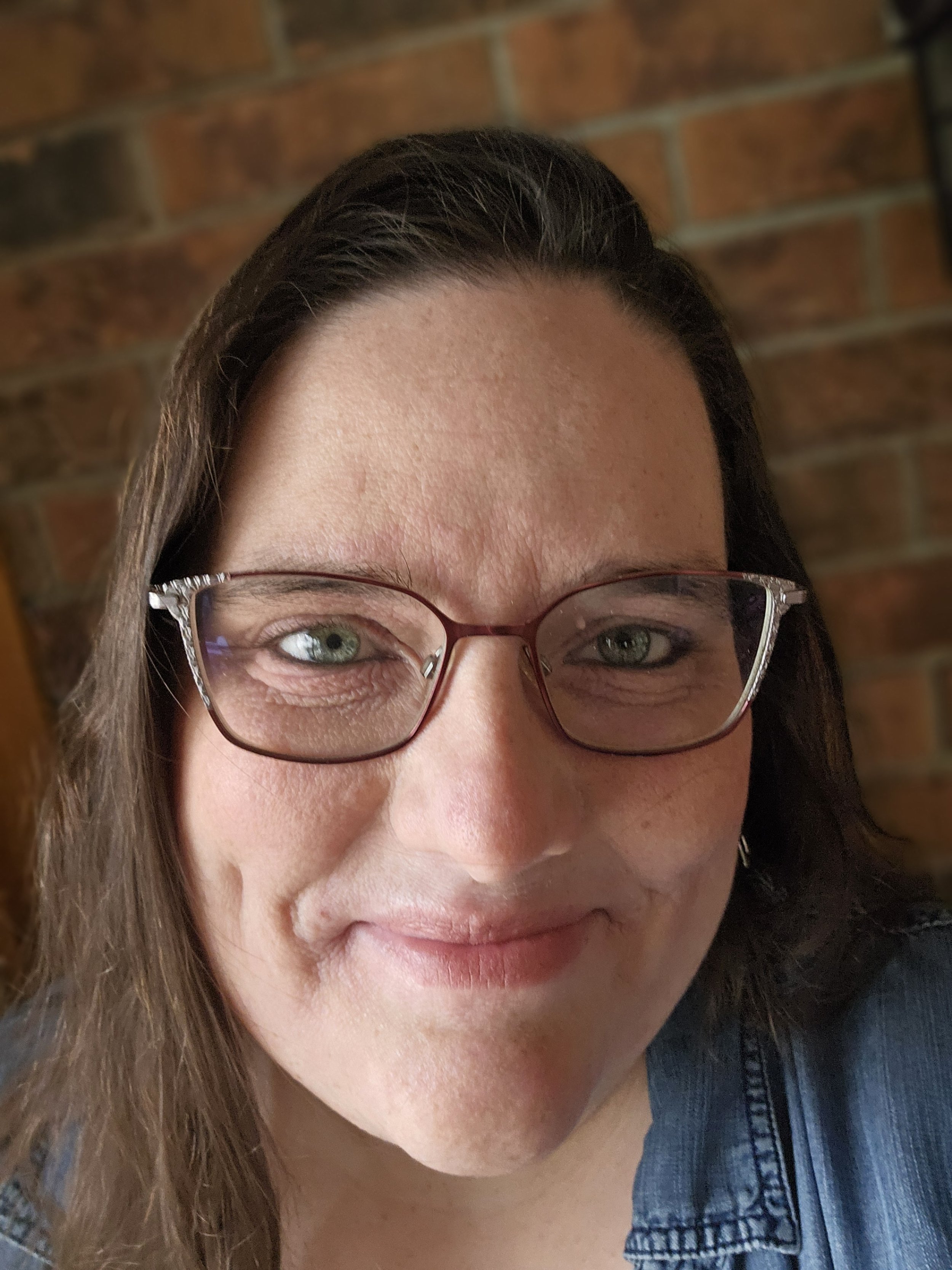 Megan Raftis smiles at the camera. Her glasses have rhinestones at the corners and her hair is down. She stands in front of a brick wall.
