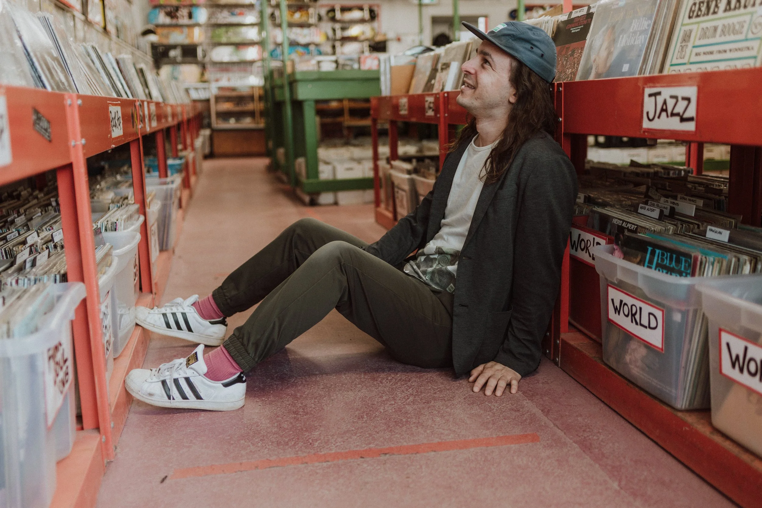 DJ Seith sits in a record store. He smiles as he relaxes  between the rows and rows of records. He wears a ball cap and his long hair is loose around his shoulders.