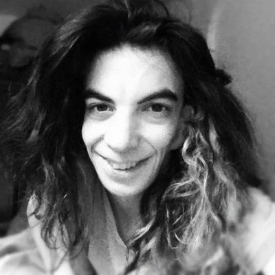 Black and white image of a white female presenting person with long curly dark hair and dark eyebrows. 