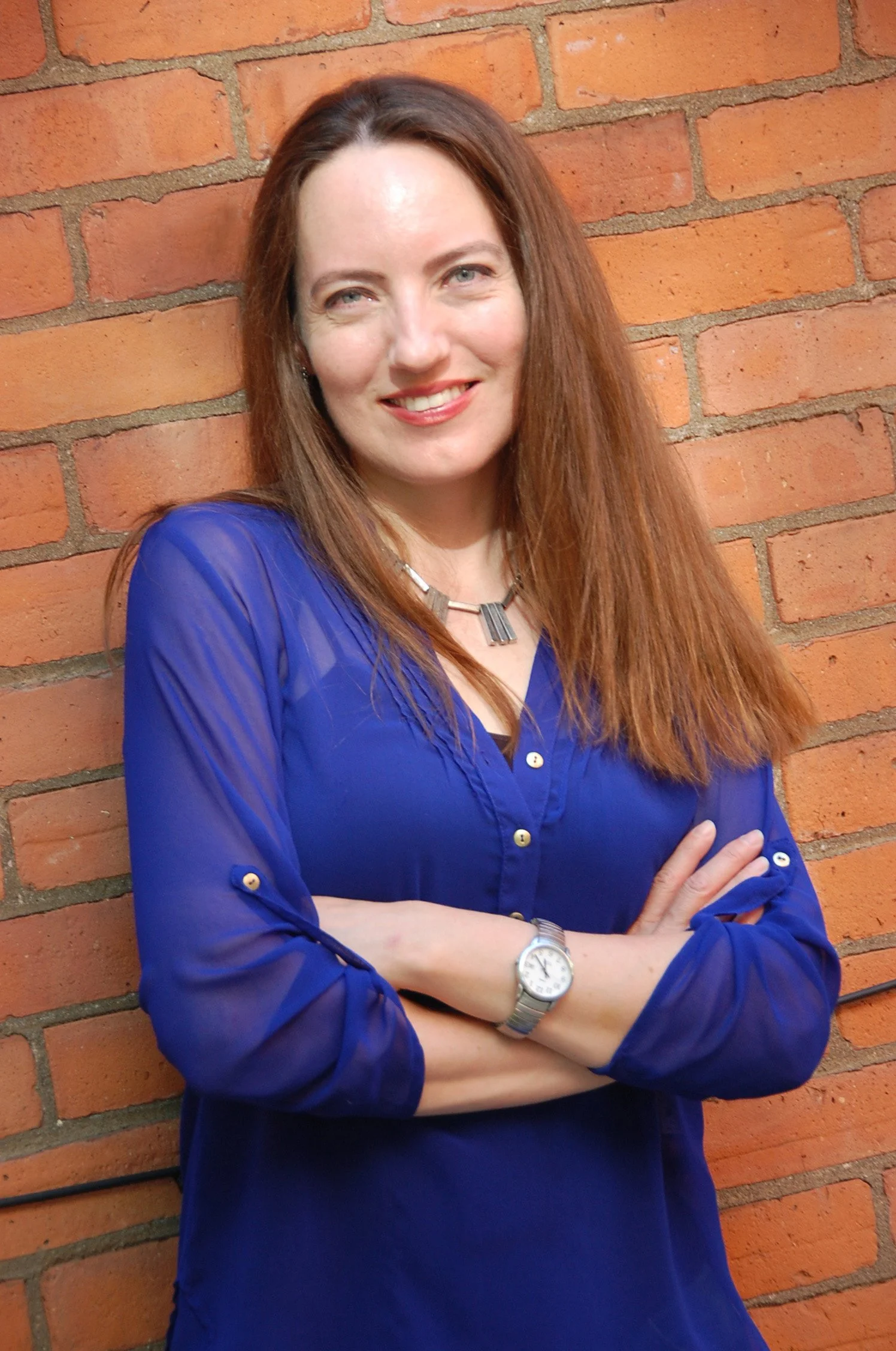 Bridget stands facing the camera, smiling, with her arms crossed easily. Her long, auburn hair is loose around her shoulders and compliments the blue buttoned shirt she is wearing.