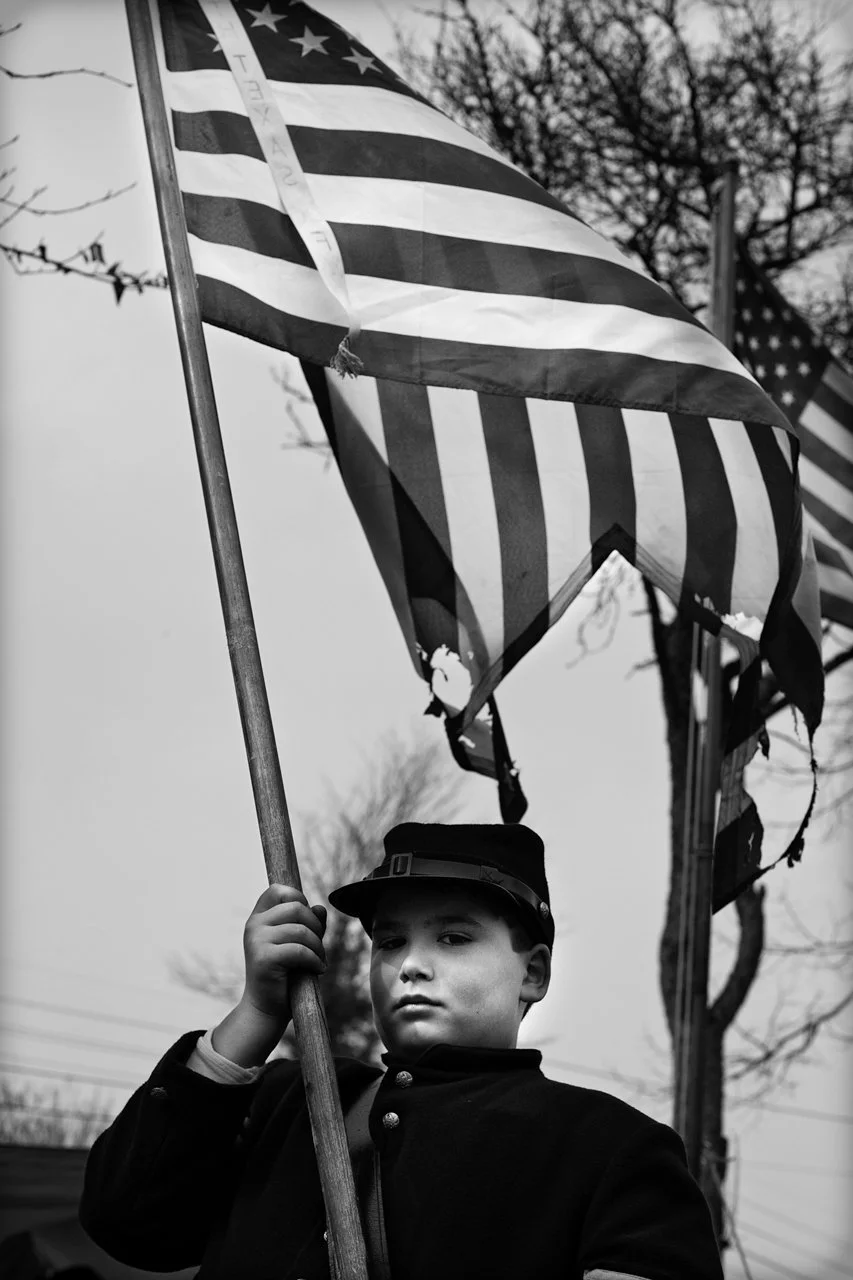 Een jongen in een militaire uniform houdt een Amerikaanse vlag vast, wat mogelijk op een herdenkings- of patriottische gebeurtenis wijst. De foto is zwart-wit en de jongen kijkt serieus naar de camera.