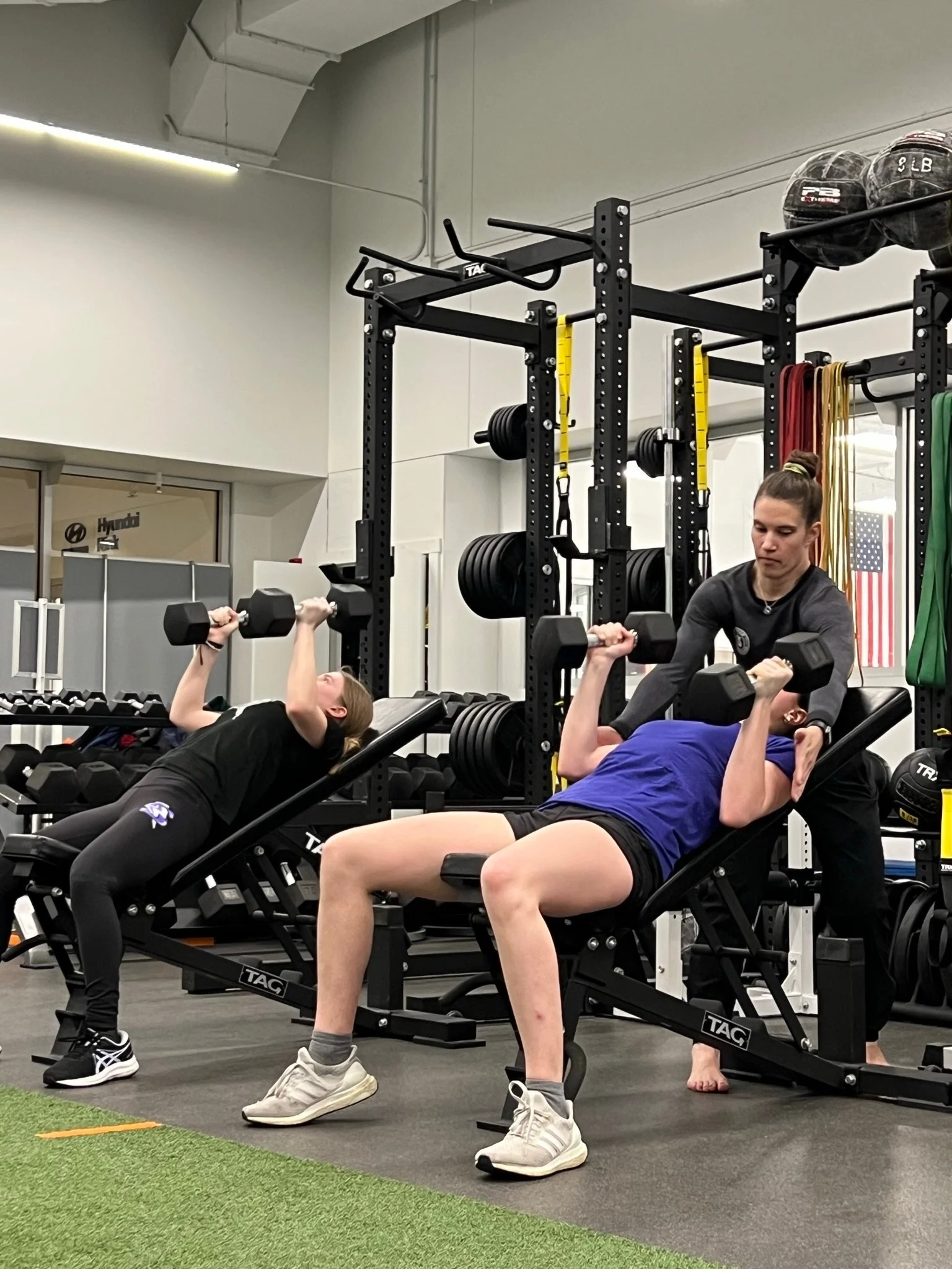 Two women are lying on adjustable benches performing dumbbell bench presses under the supervision of a trainer in a gym. The gym has black weight racks, medicine balls, resistance bands, and American flags in the background.