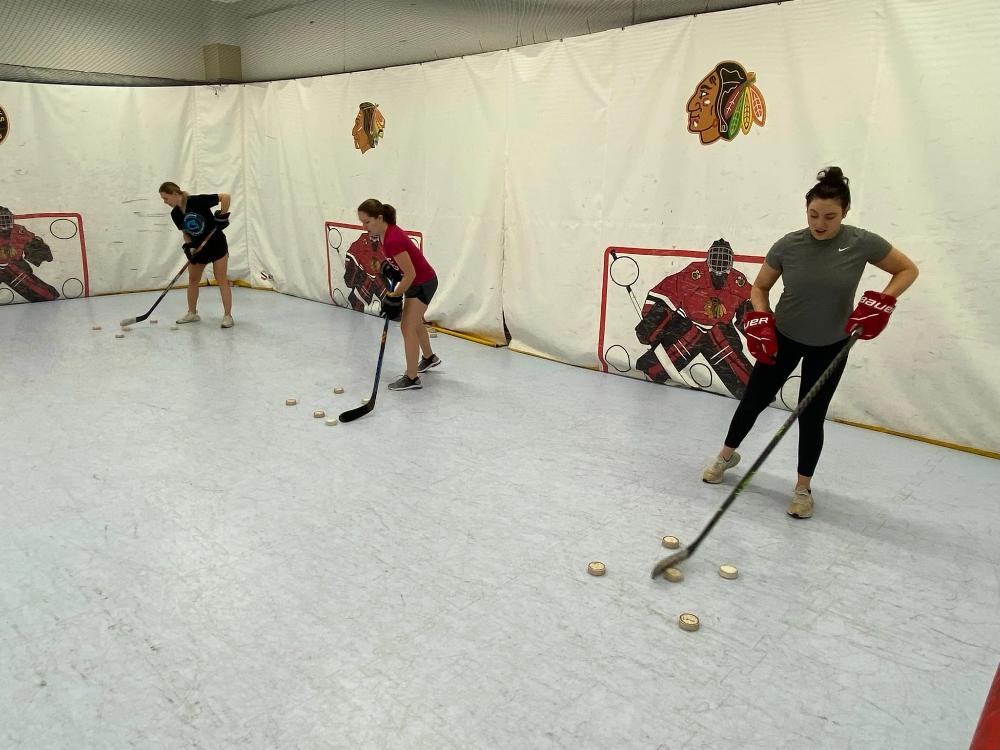 Three women practicing ice hockey skills on an indoor rink, with one woman in a gray shirt and red gloves in the foreground, and the other two women in black and pink shirts behind her, all using hockey sticks to hit pucks on the ice.