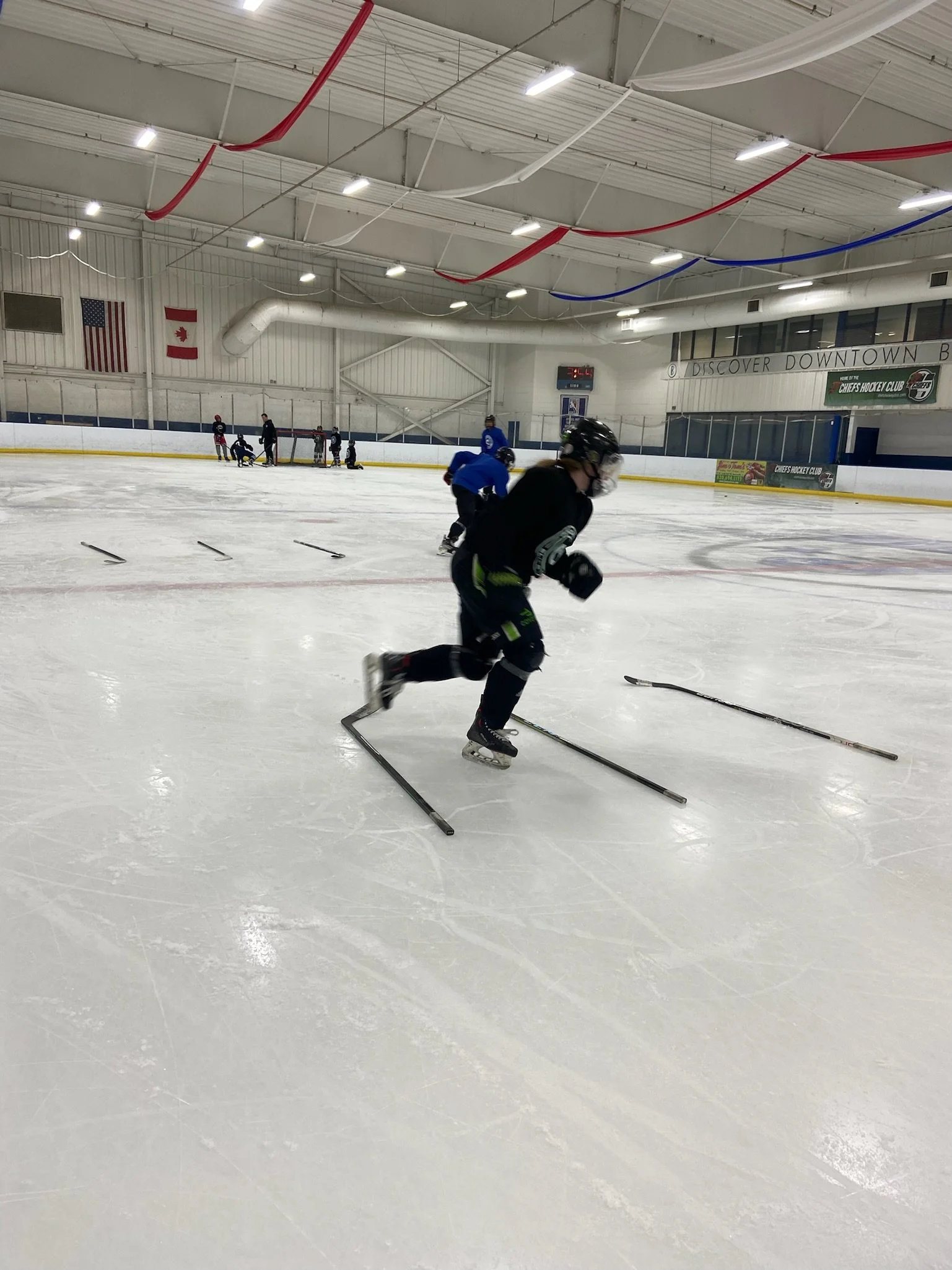Ice hockey players practicing drills on an indoor rink, with some players skating and others positioned near the boards, flags on the wall, and a scoreboard in the background.
