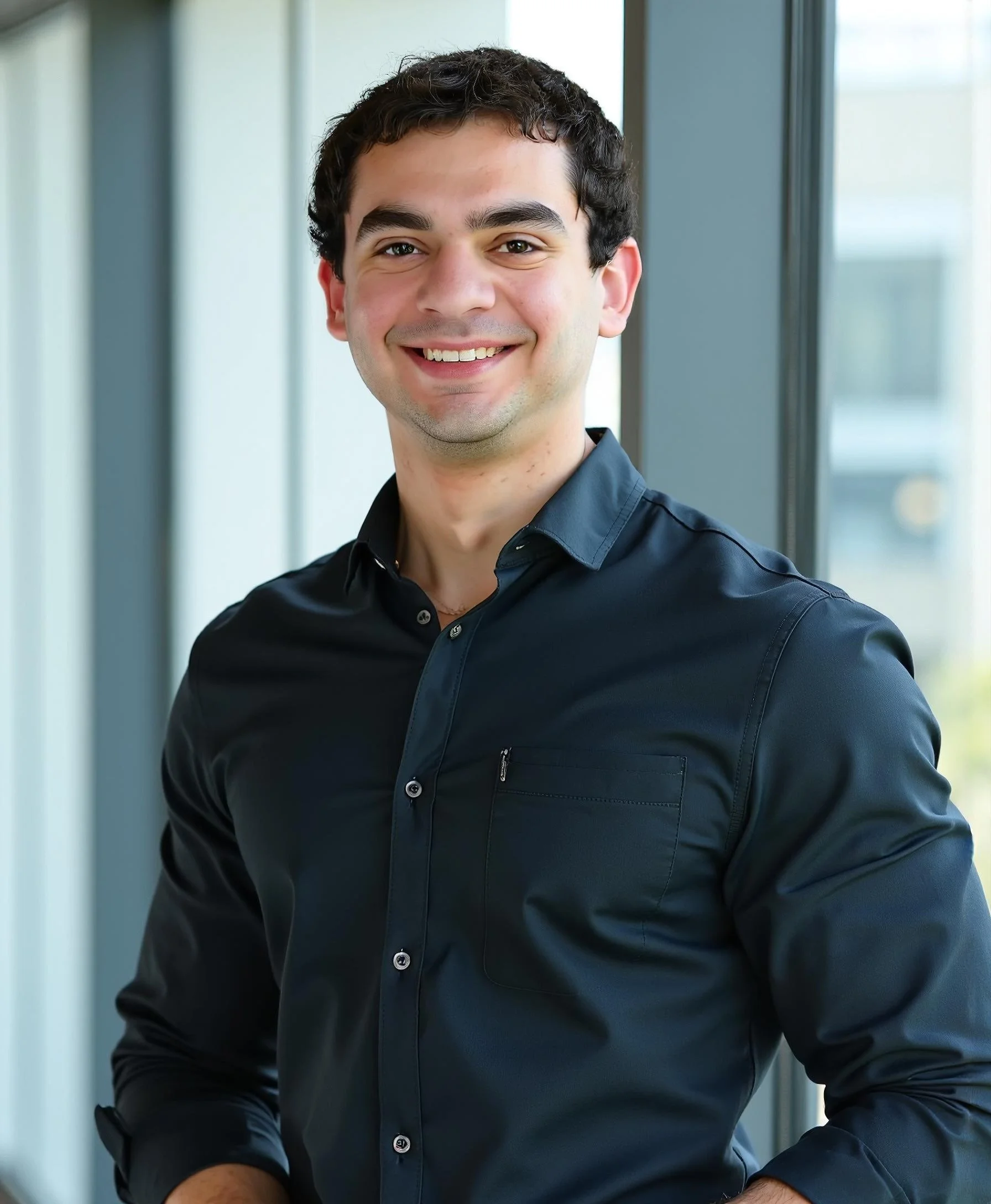 A smiling man in a black button-up shirt standing in front of windows in an office setting.