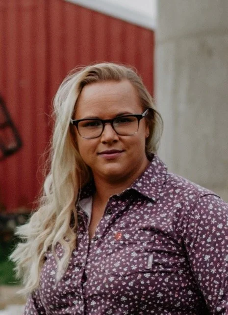 A woman with long blonde hair, glasses, and a patterned purple shirt, standing outdoors near a red building and a concrete wall.