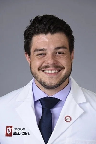 Male doctor in a white lab coat with the Indiana University School of Medicine logo, smiling against a gray background.