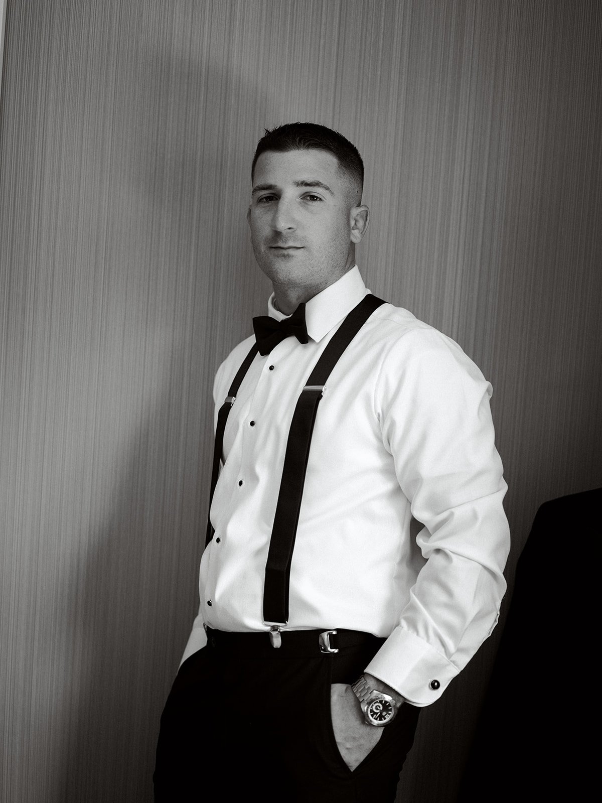 Grooms leans against a wall, looking at the camera