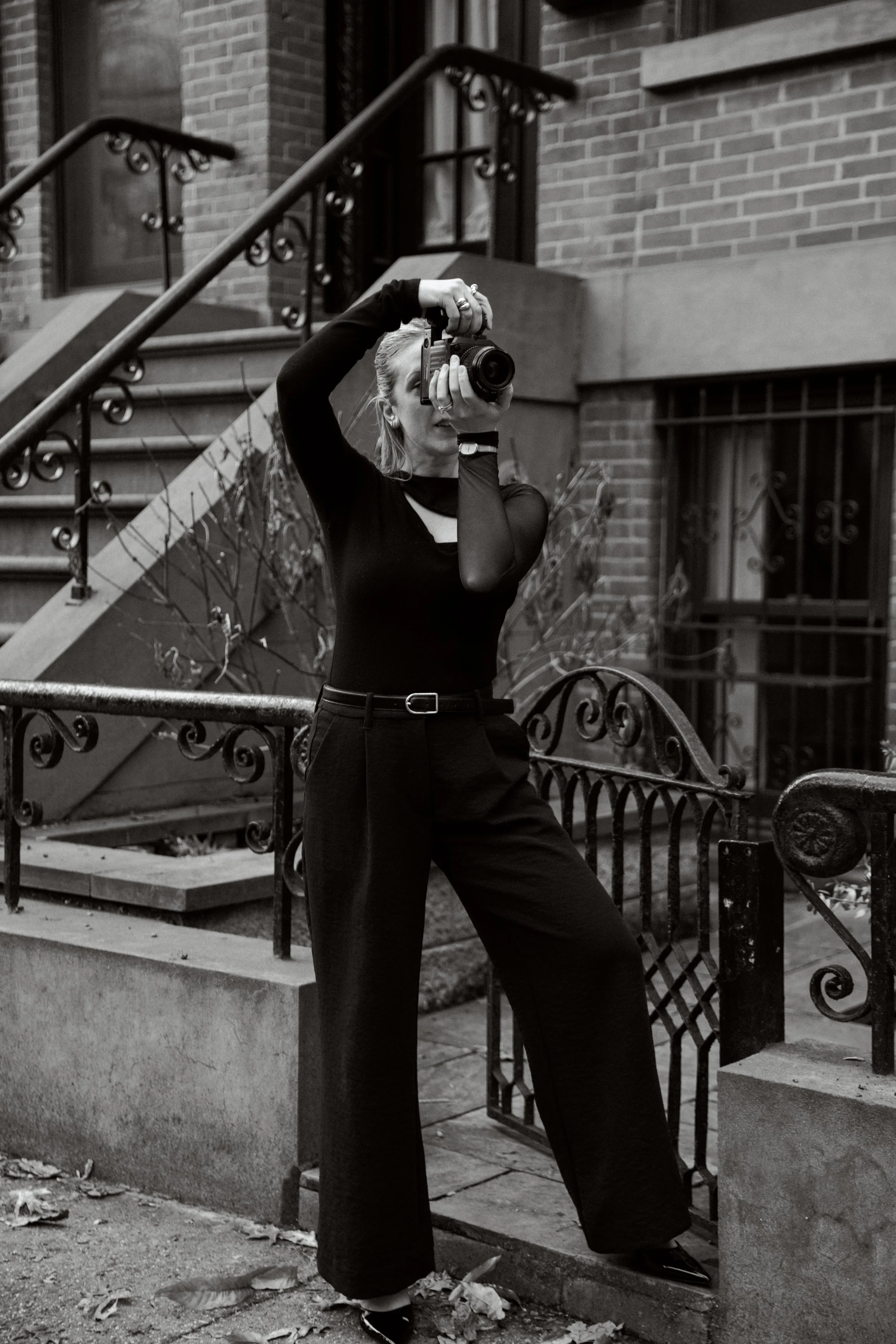 A woman taking a photograph with a camera outdoors in front of a residential building with stairs and iron railings. She is dressed in black, standing on a sidewalk among fallen leaves.