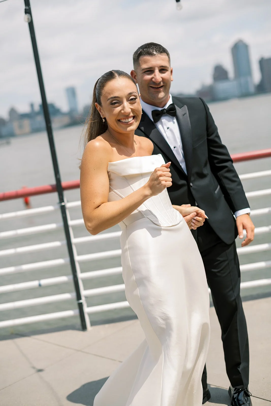 bride and groom smiling at camera, walking along pier with NYC skyline in the background. Battello wedding
