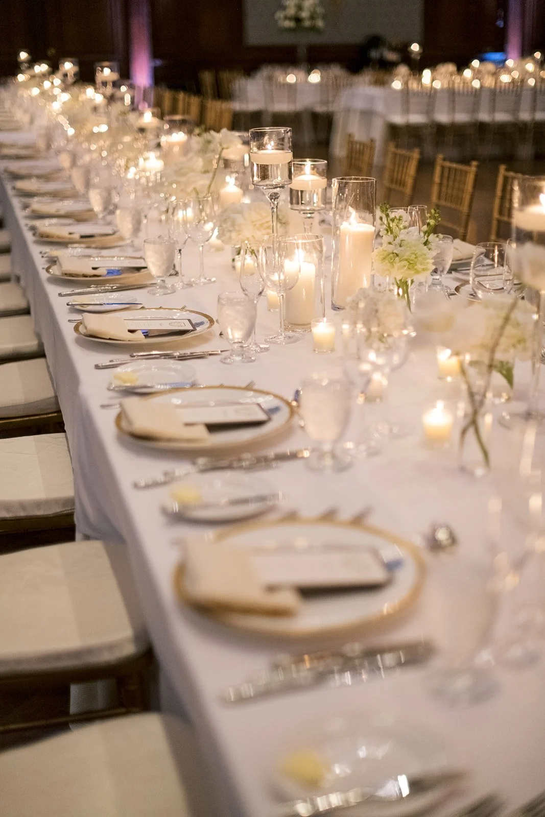 Long table set for wedding reception with white and gold chargers, white menus, votive candles and single white flowers in small vases. Union League of Philadelphia