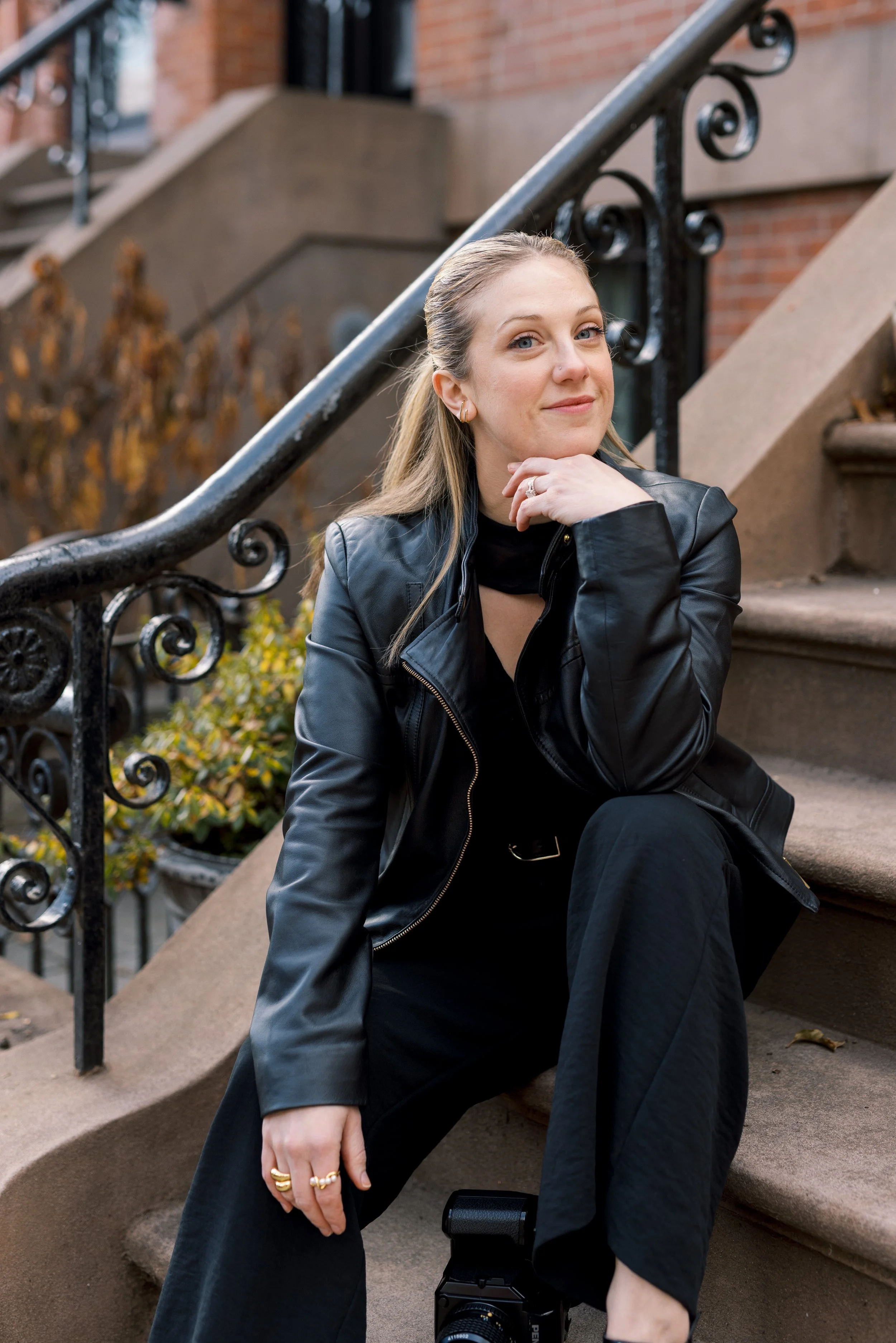 A woman sitting on brownstone stairs in an urban neighborhood, wearing a black leather jacket and black pants, with a camera on the step below her.