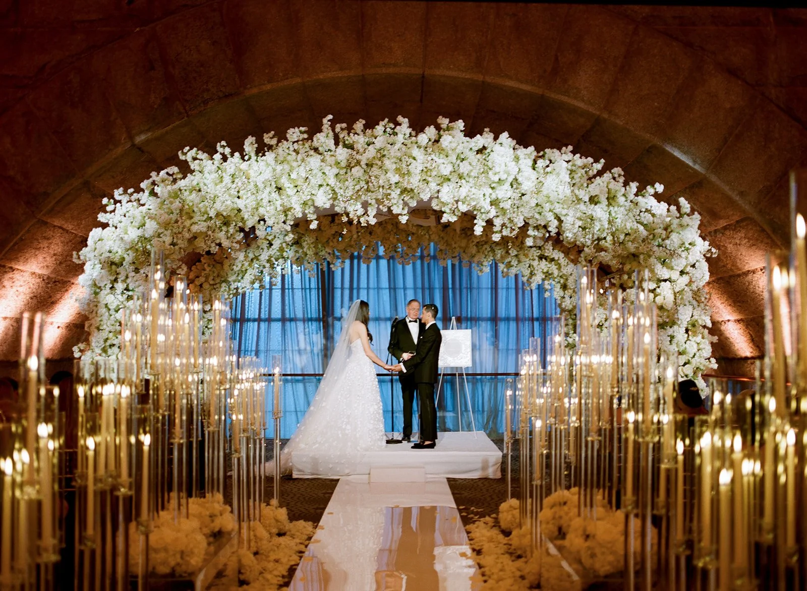 bride and groom under large chuppah with floral arch of white flowers and candelabras lining the ceremony aisle. film photo.