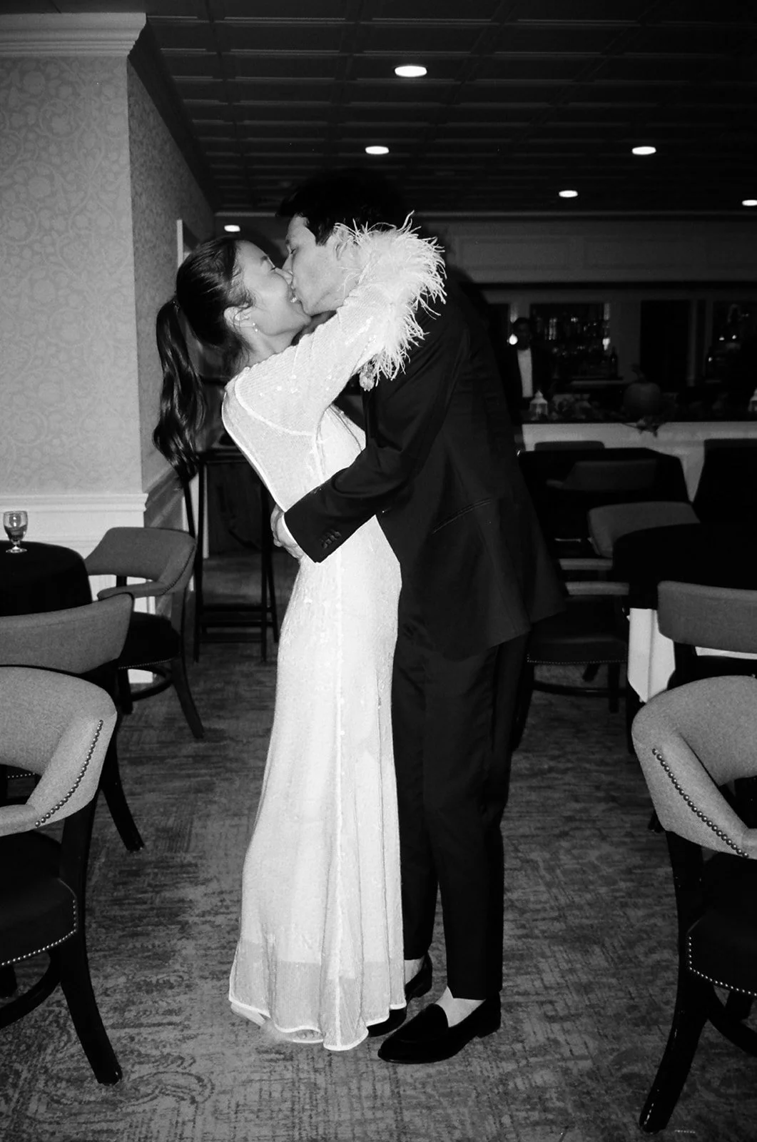 bride and groom kissing. Bride is wearing a sequin long dress with feather cuffs and groom is in a black tuxedo. Black and white photo