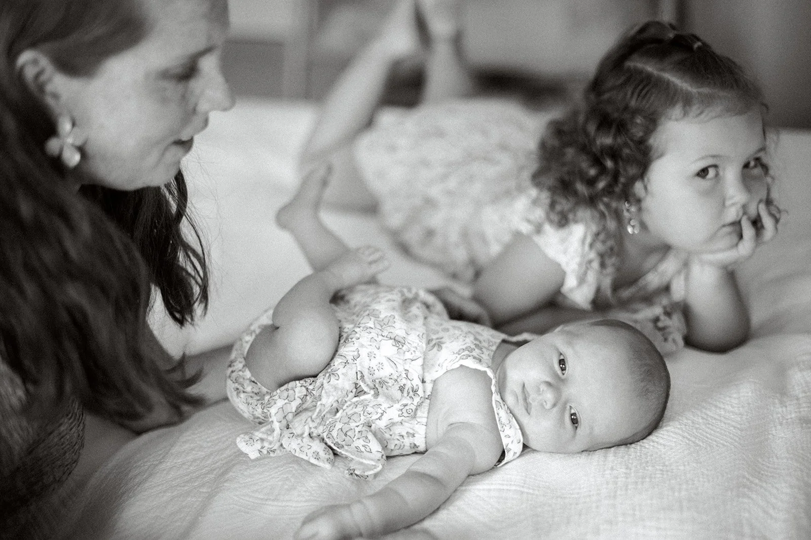 Mom leans over newborn daughter as older sibling looks at the camera in the background