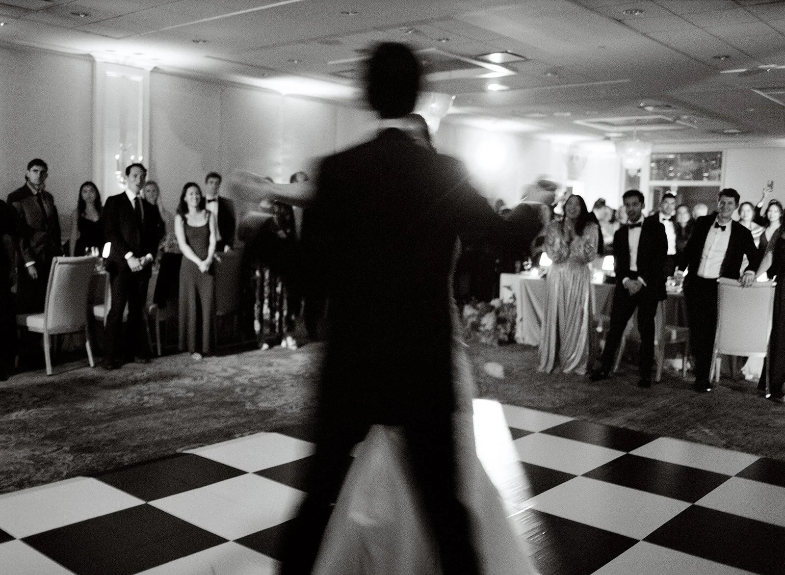 blurred image of bride and groom dancing in the foreground with guests in focus in the background. Checkered dance floor