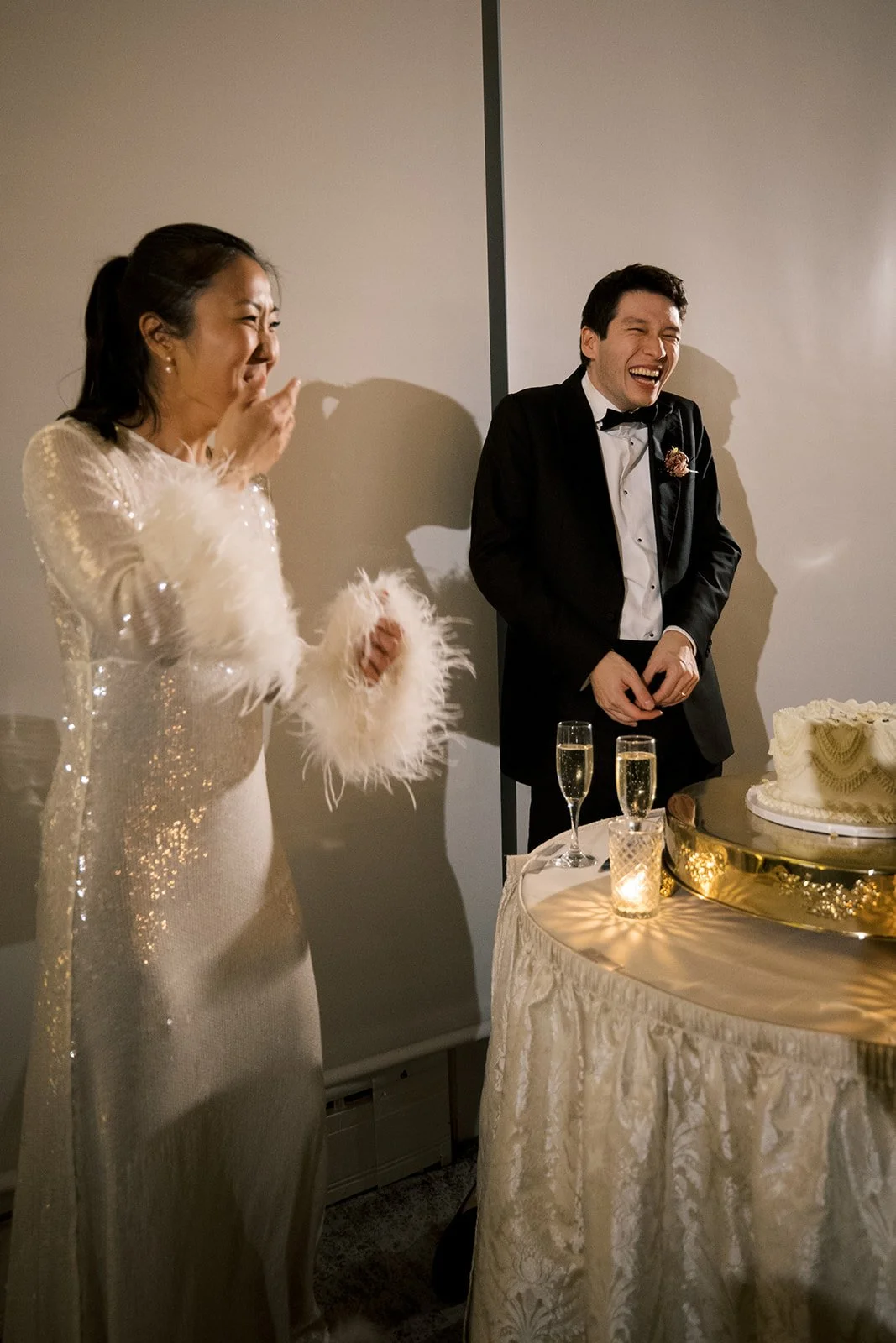 Bride wears sequin dress with feather cuffs and covers her mouth as she and the groom laugh together standing next to their wedding cake