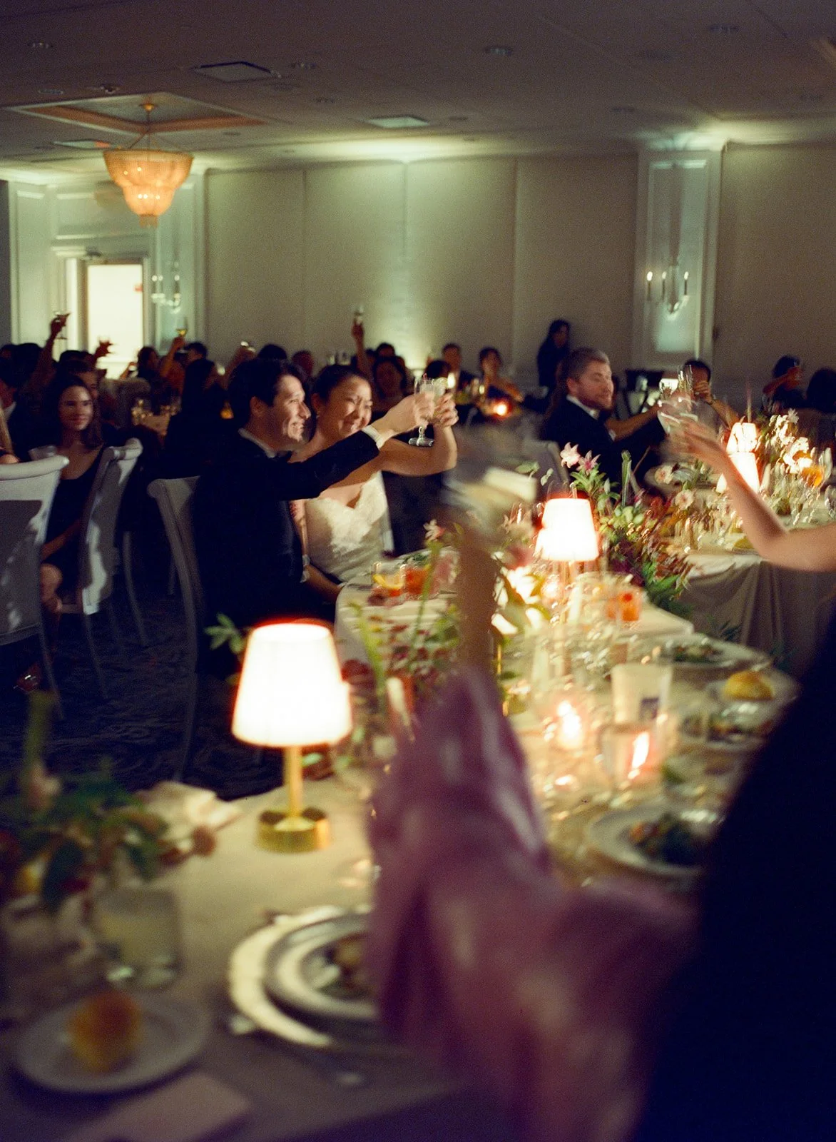 wedding guests and bride and groom toasting glasses as they sit at a serpentine wedding table with tabletop lamps