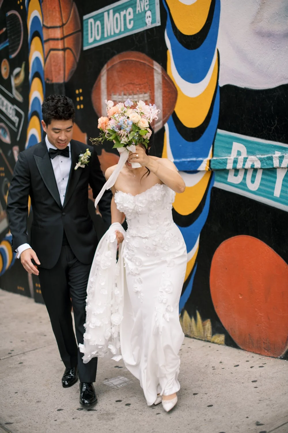 Bride and Groom on the sidewalk in Brooklyn infront of a large colorful mural. Bride holds bouquet above her head to shield from rain