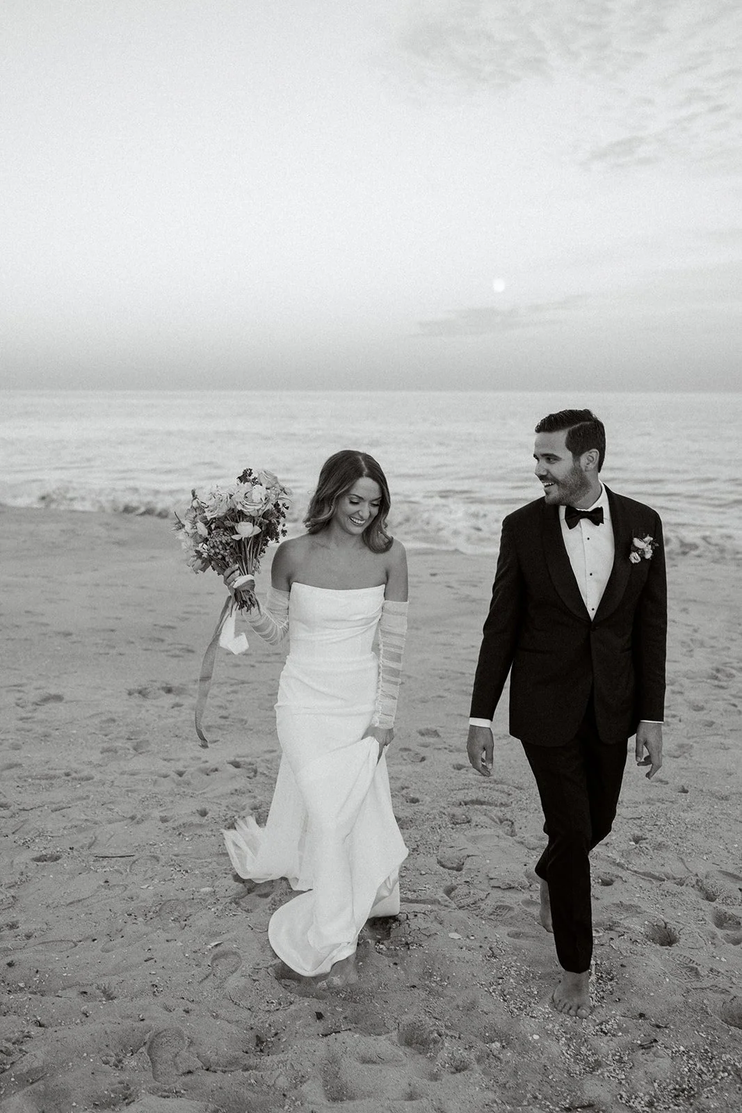 black and white portrait of bride and groom walking on the beach with the ocean in the background