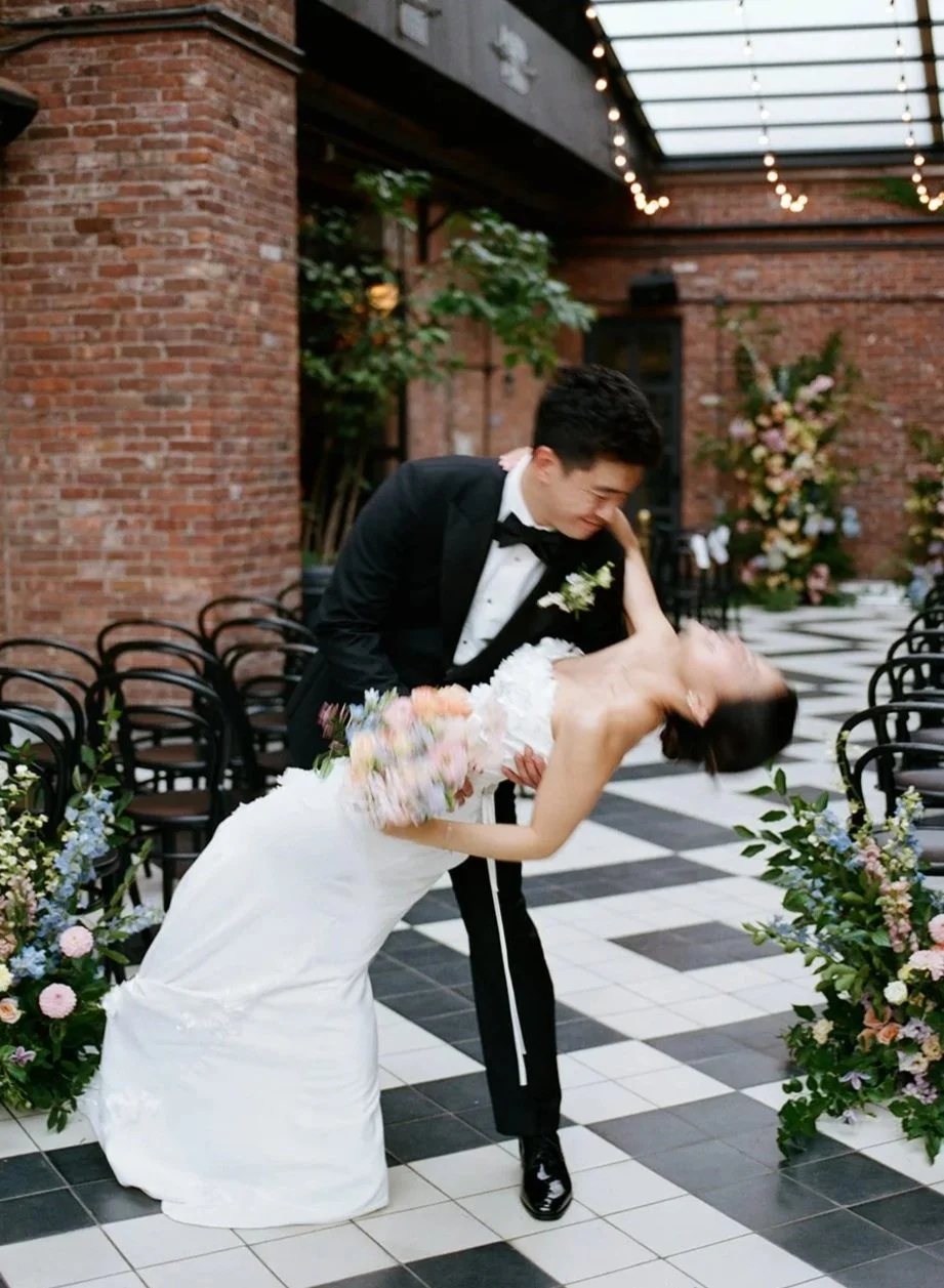 A groom in a black tuxedo dips a bride in a wedding dress holding a bouquet of flowers at a wedding venue with brick walls, string lights, and floral decorations.