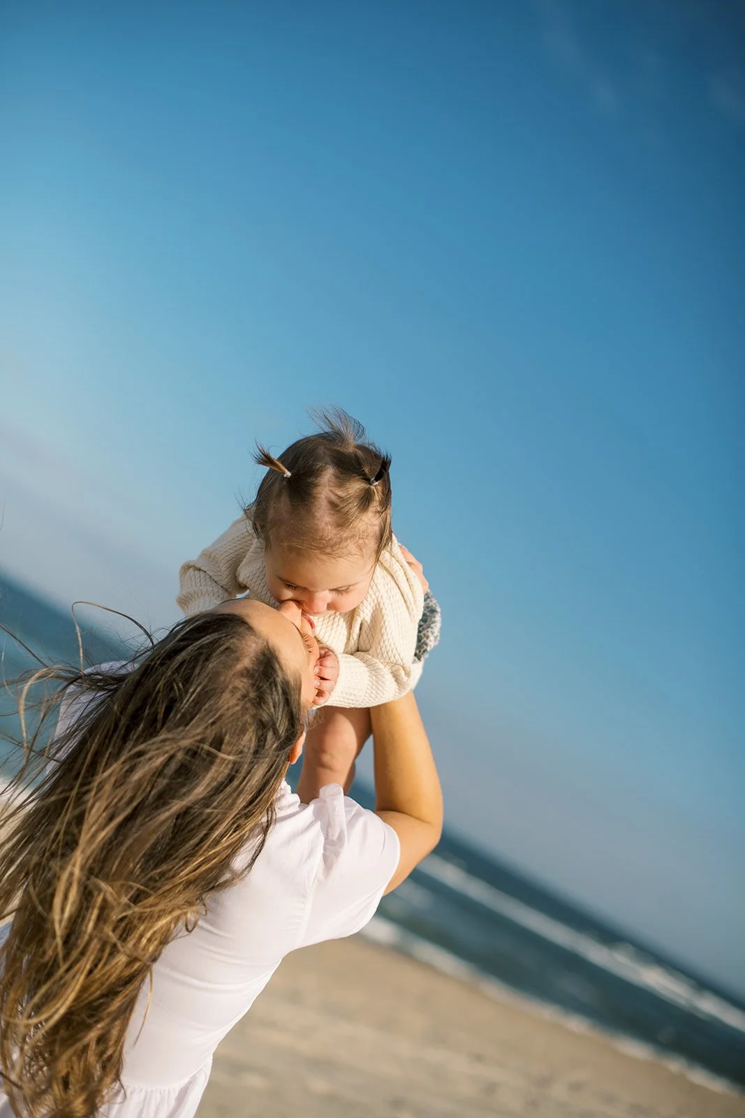 Mother holds daughter above her head at the beach as she kisses her cheek