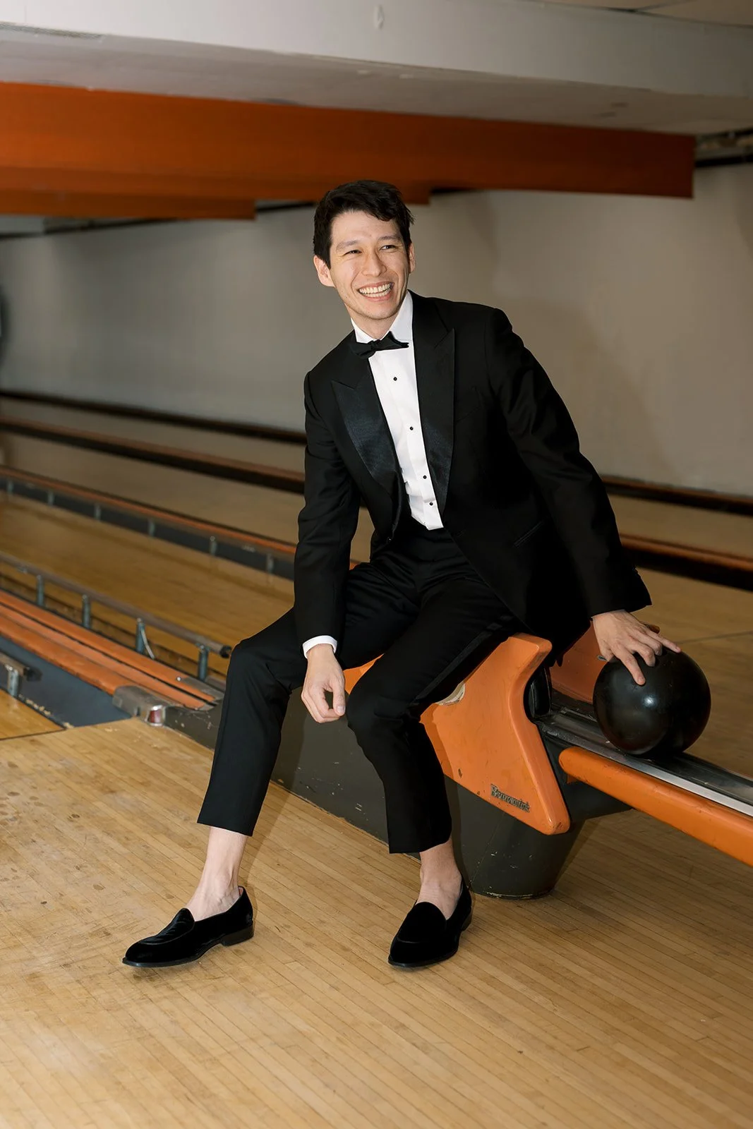 Groom sitting on bowling alley ball rack smiling at camera wearing a black tuxedo