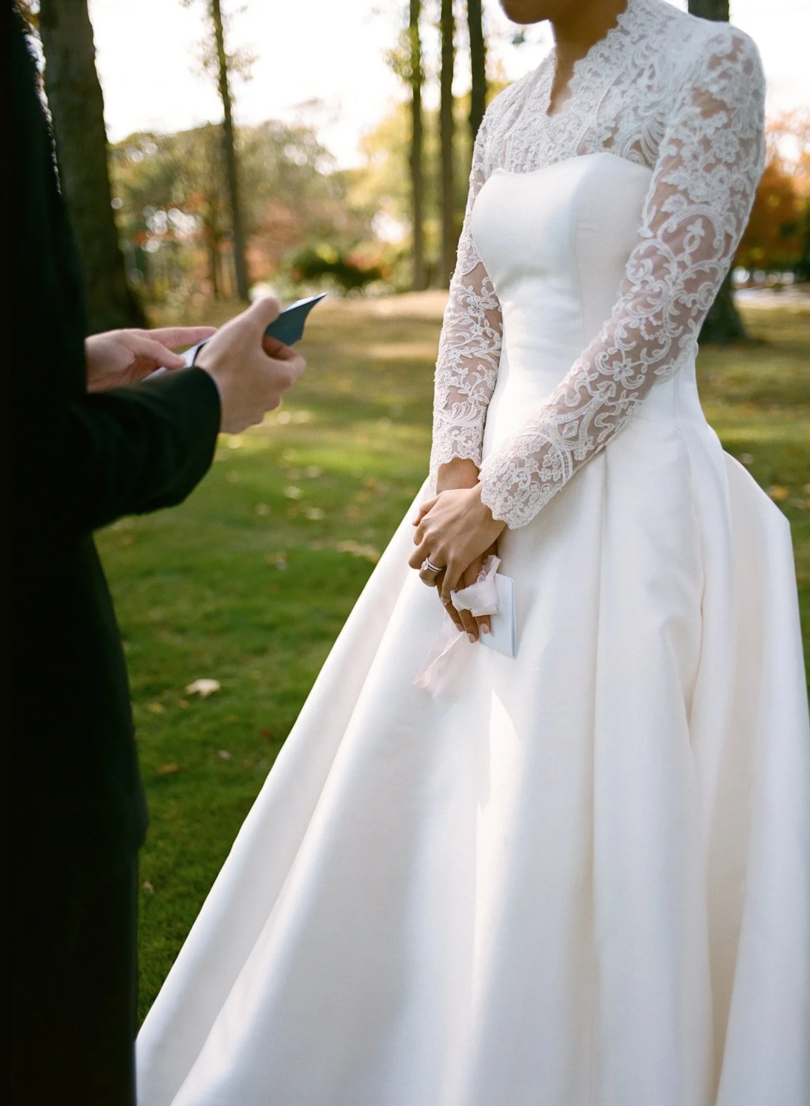 Cropped image of bride's hands folded together against her white dress with lace sleeves. Groom's hands holding vow book can be seen on the side