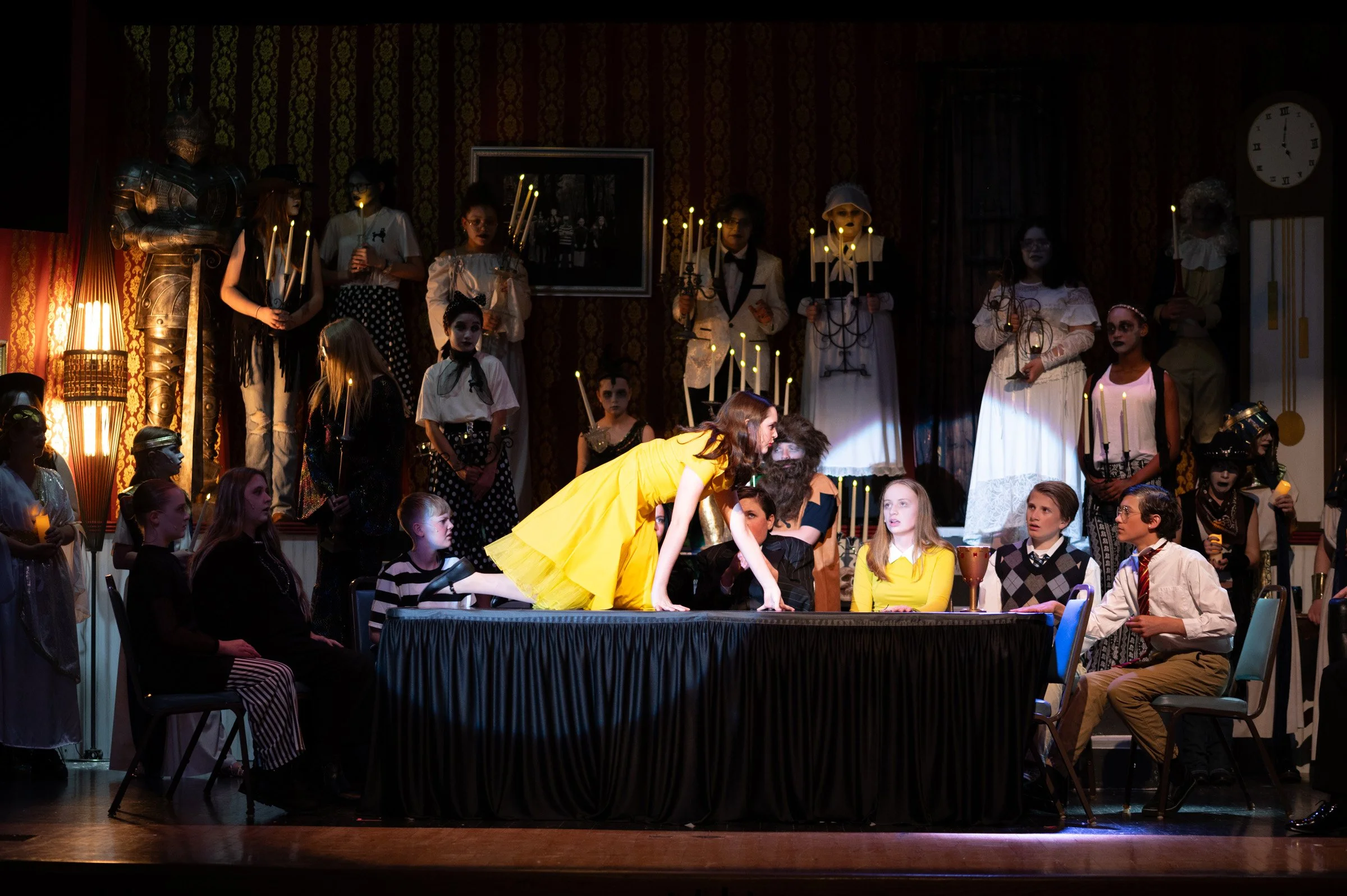 A theatrical stage scene with actors and children, some dressed in costumes, and a woman in a bright yellow dress leaning over a table with a bearded man. The backdrop is dark with patterned wallpaper, candles, and a clock.