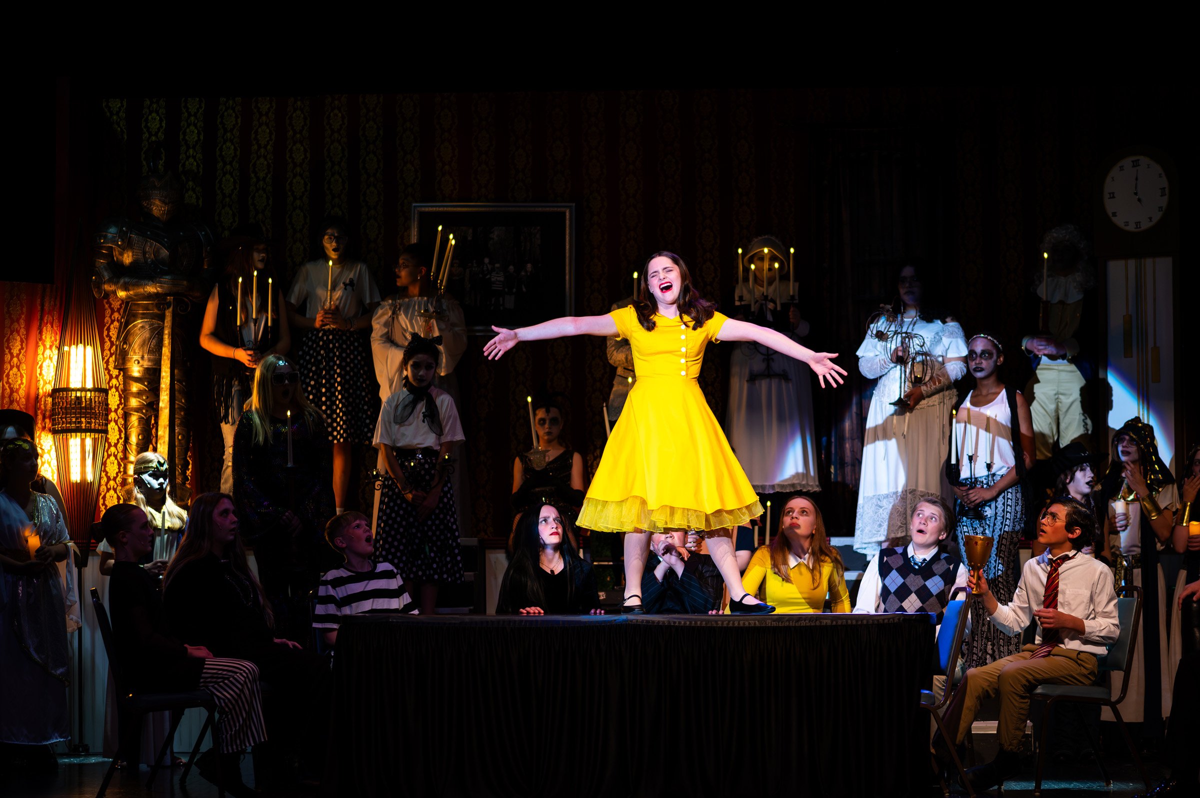 A woman in a bright yellow dress standing on a table with arms wide open, surrounded by children and adults in costumes on a stage during a theatrical performance.