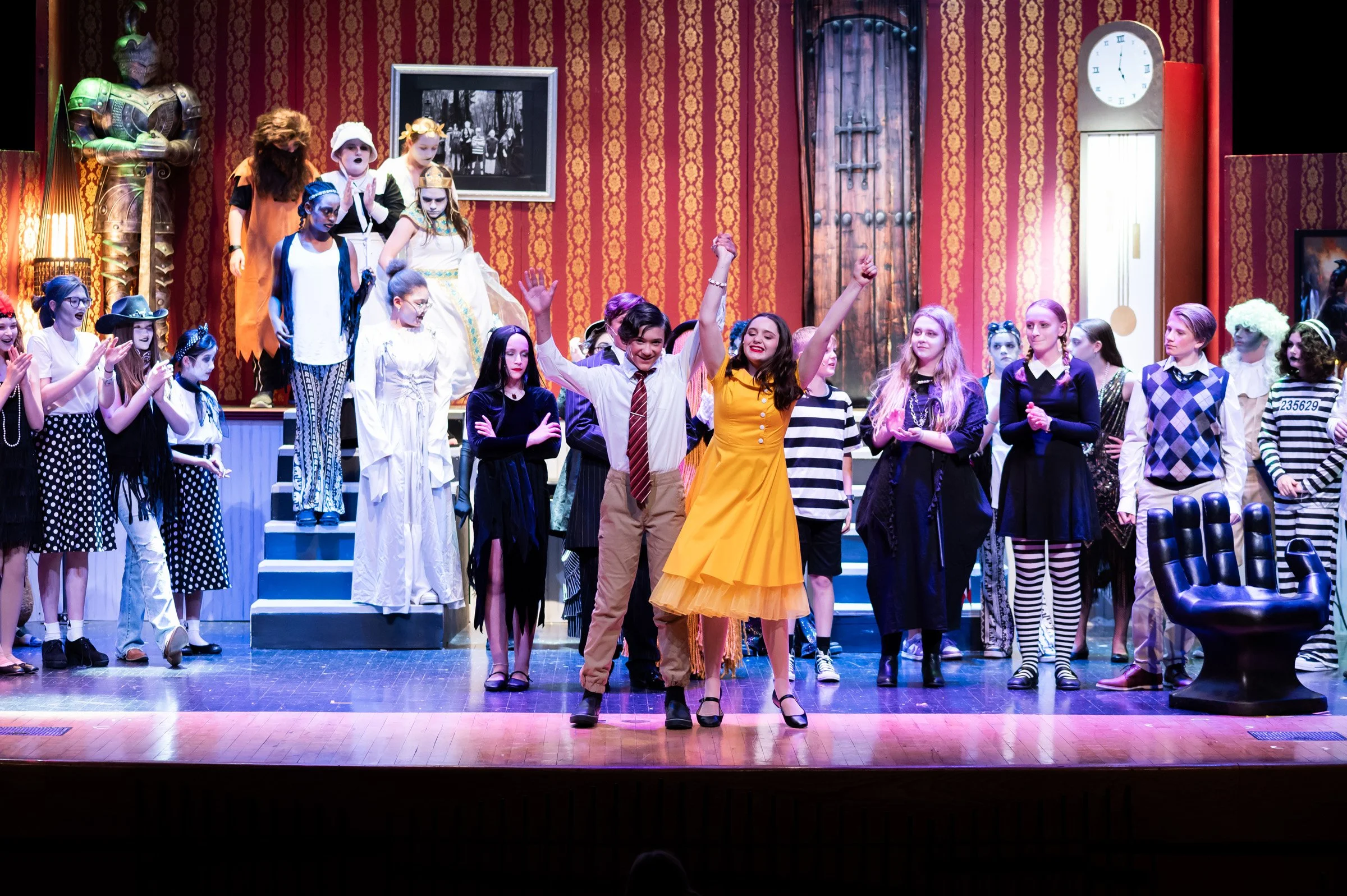 The cast of a theatrical play on stage, with two actors in the center holding hands and smiling, surrounded by other cast members in costumes, all clapping and celebrating.