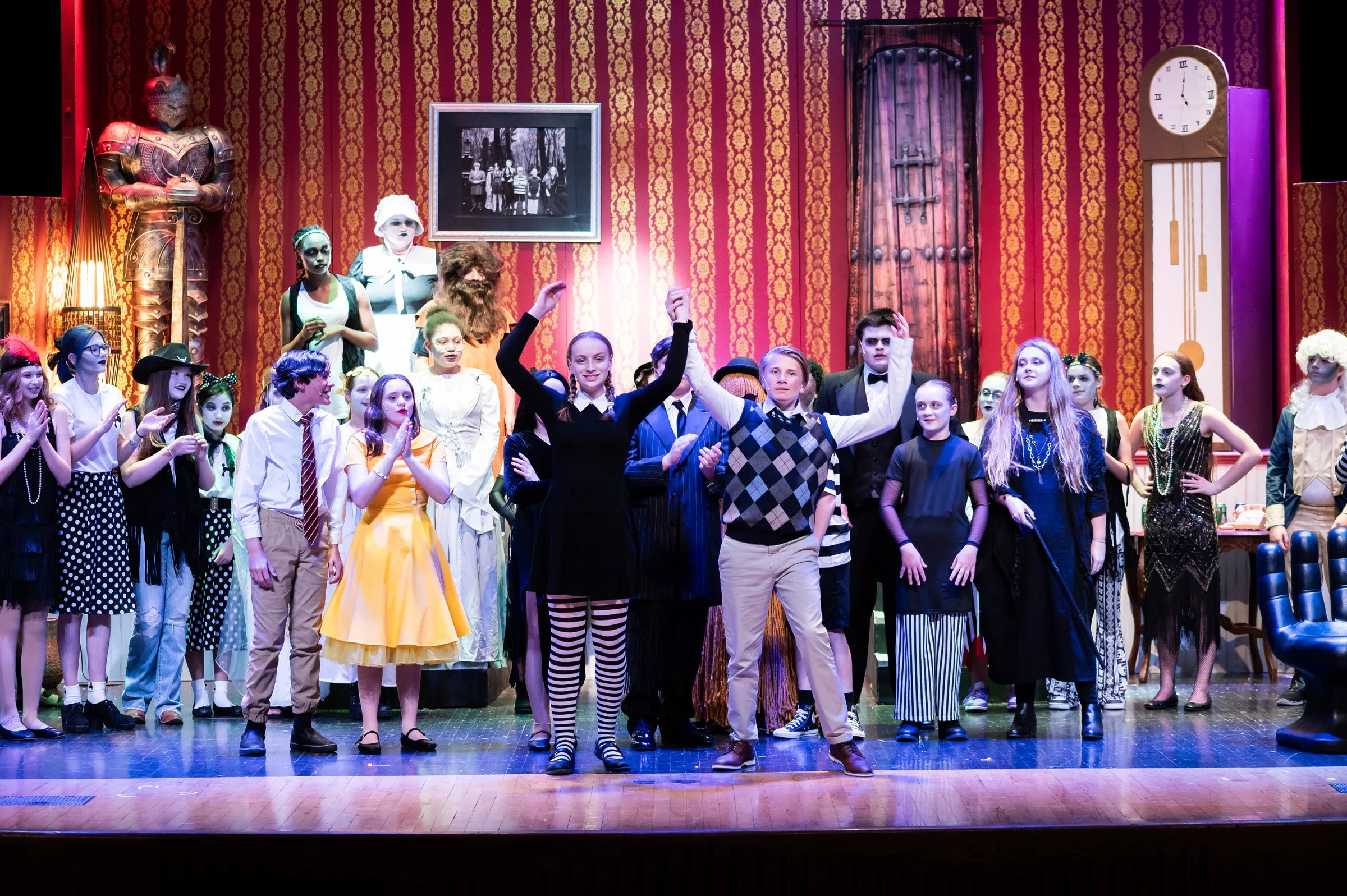 A group of children and teenagers dressed in costumes on a stage, possibly after a performance, with a red patterned wallpaper background, a large cabinet, and a clock on the wall.