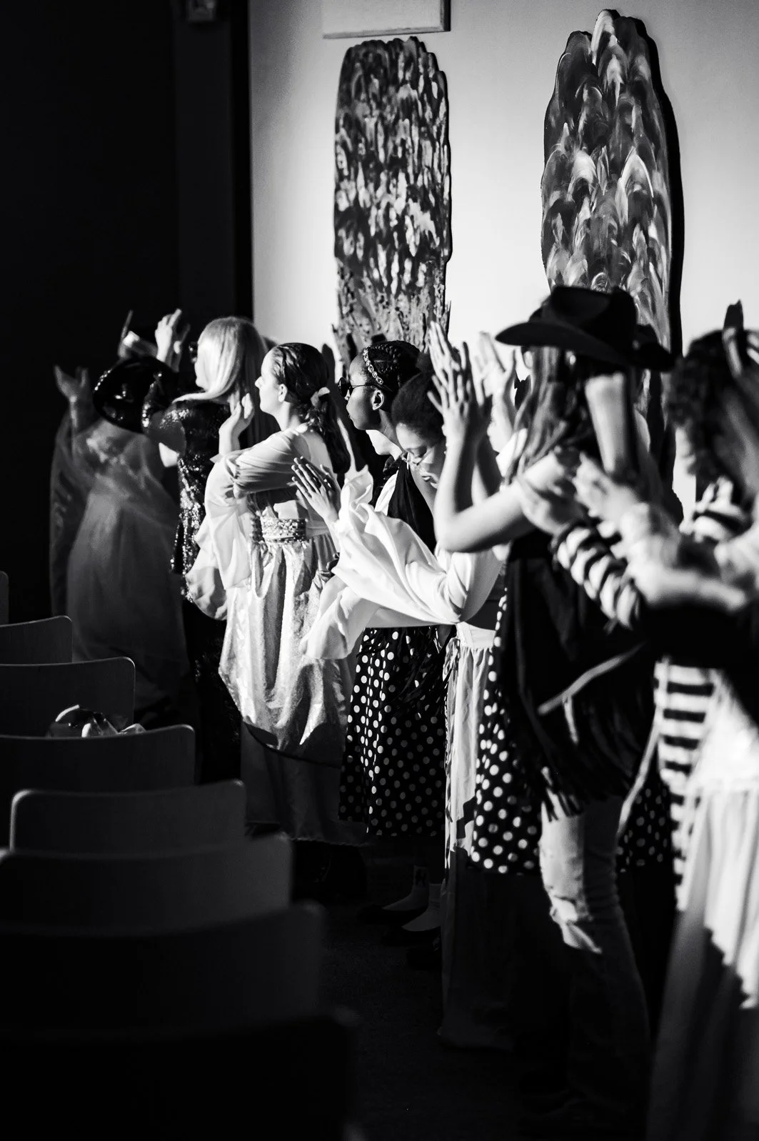 A group of people standing on stage during a performance or event, dressed in various costumes, with some using their hands or clapping, in front of abstract artwork on the wall, in black and white.