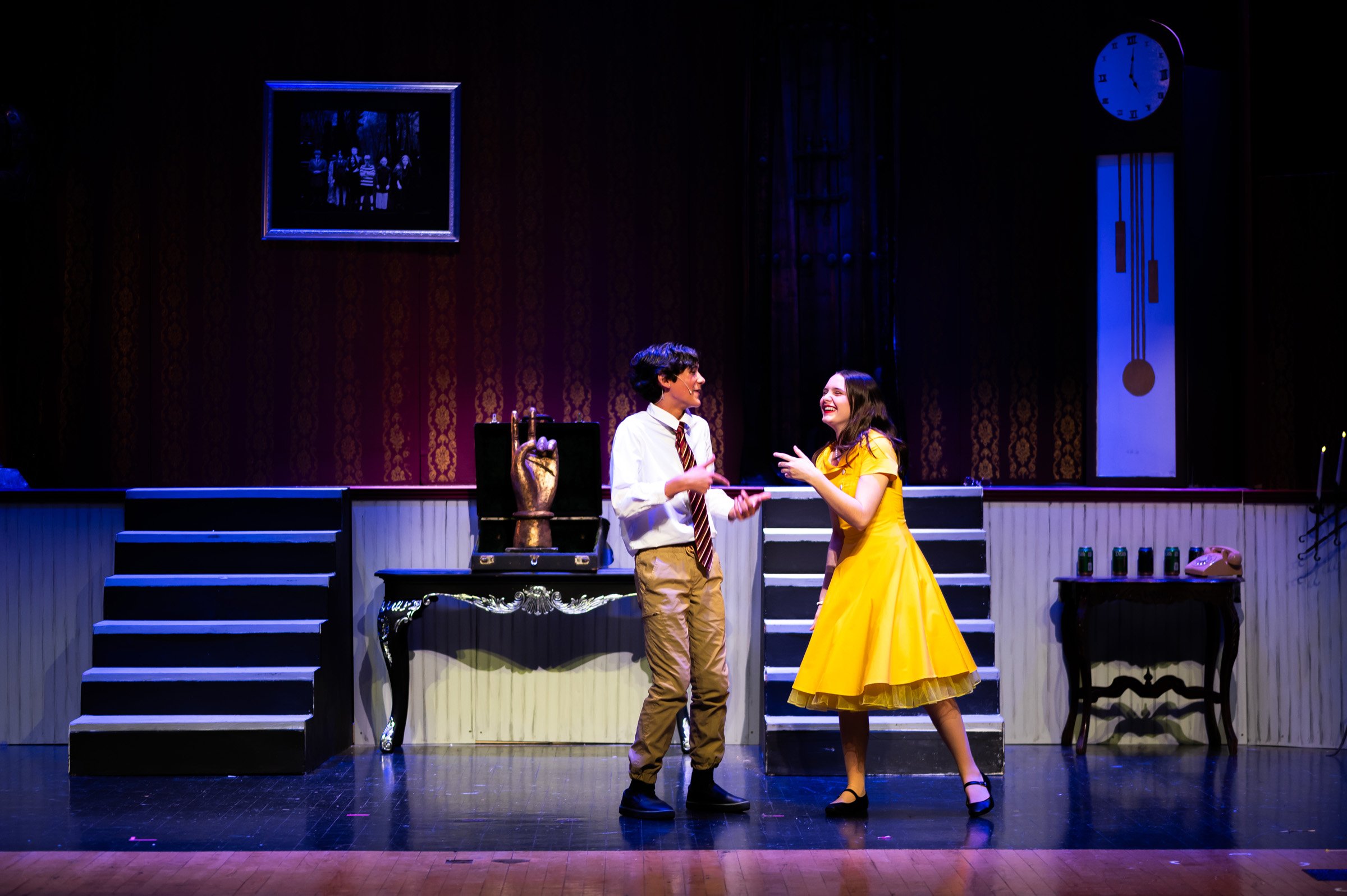 Two actors on stage in a theater, a man in a white shirt and striped tie and a woman in a yellow dress, appear to be singing or talking, with the woman laughing. Stage set includes stairs, a table, and a clock in the background.