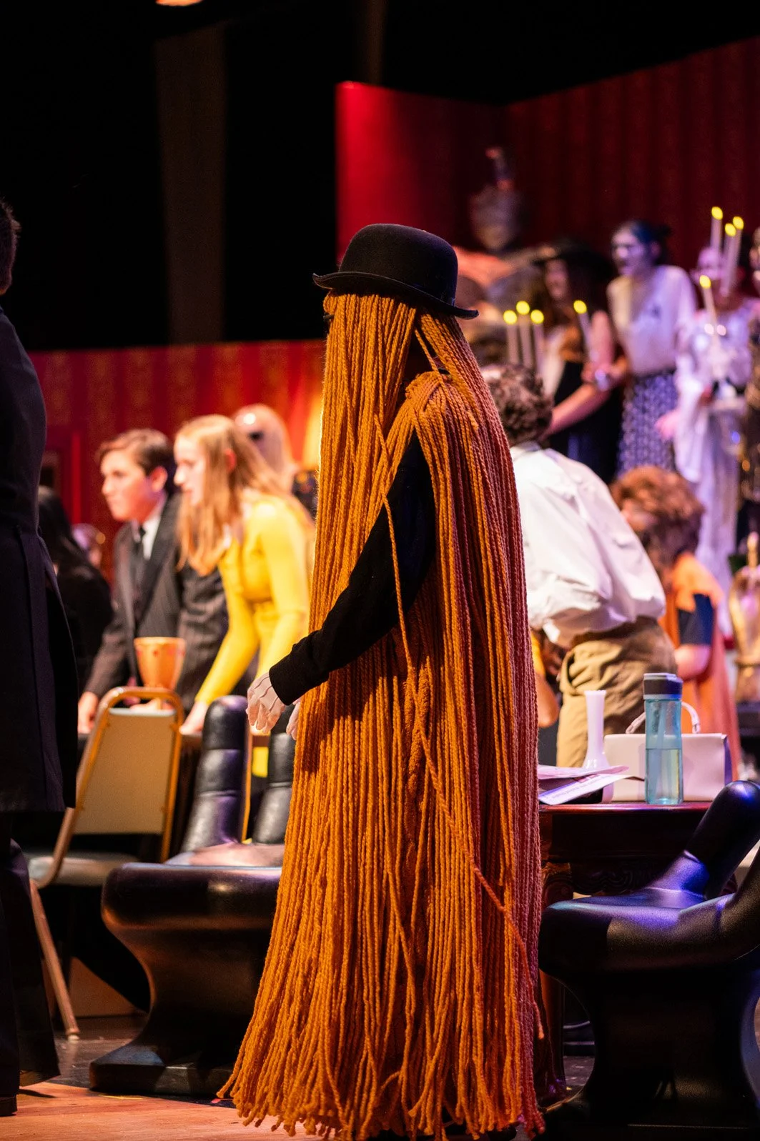 Person with long, orange dreadlocks and a black hat standing in a crowded room with people and red curtains in the background.