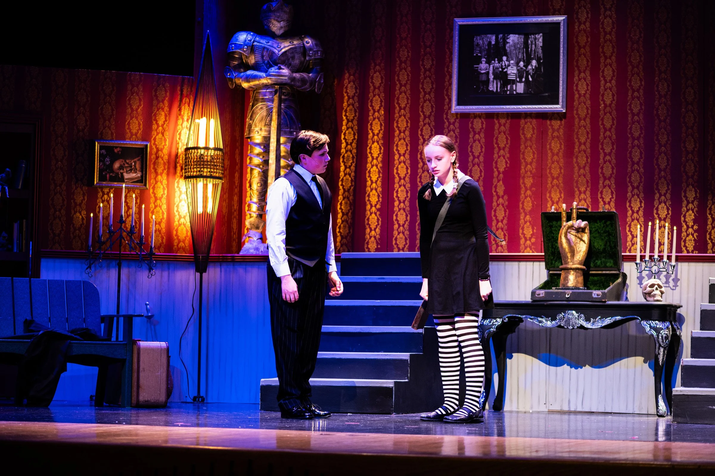 Two actors on stage in a theater set, with a red and gold patterned wallpaper background, a large knight armor statue, and vintage furniture, including a table with a golden hand sculpture, a skull, candles, and framed photos. The actors are facing e