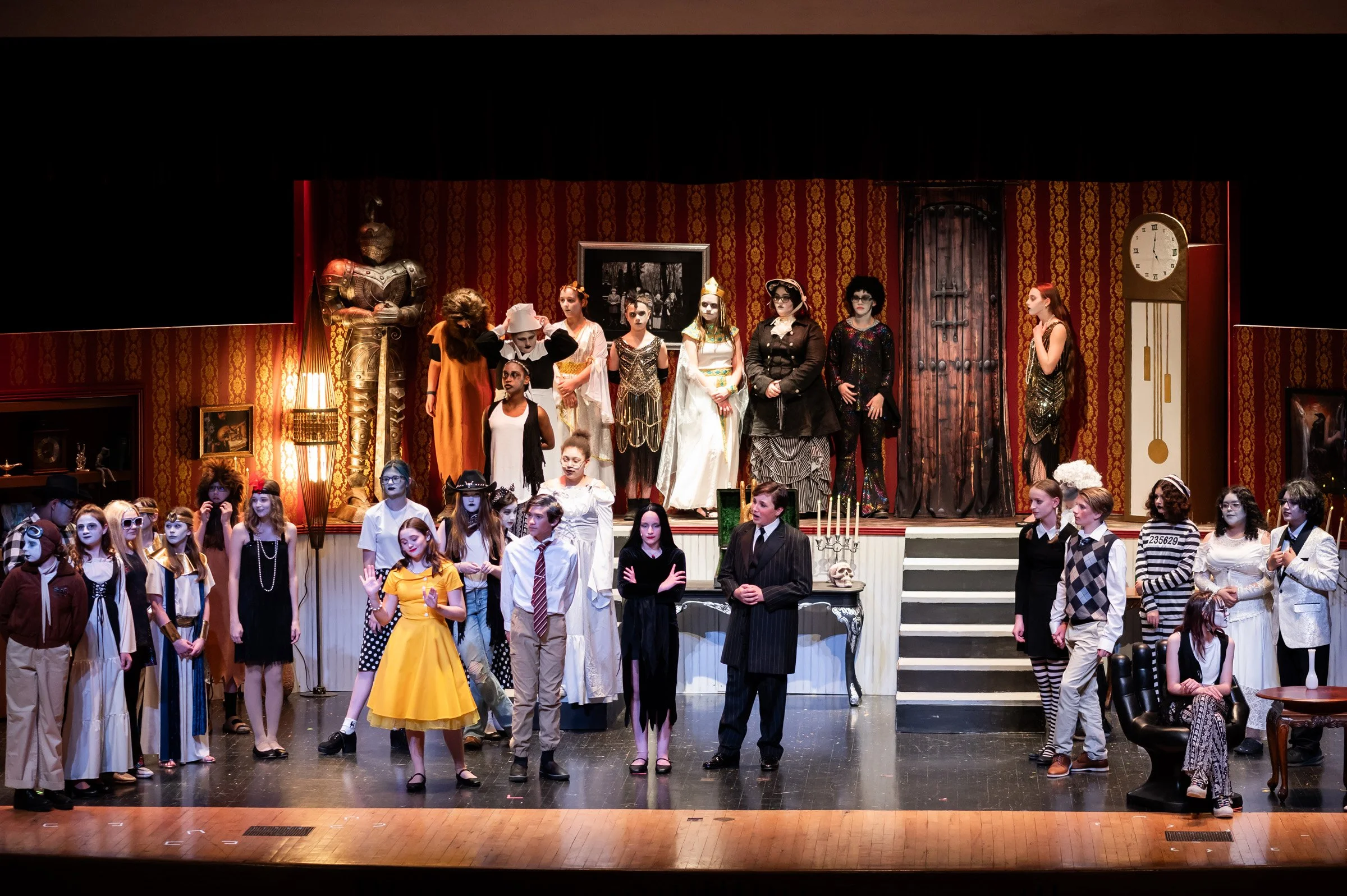 The cast of a theatrical play on stage, dressed in various costumes, with some standing on stairs and others on the main stage area, set against a vintage, ornate background with red wallpaper, a large clock, and various props.