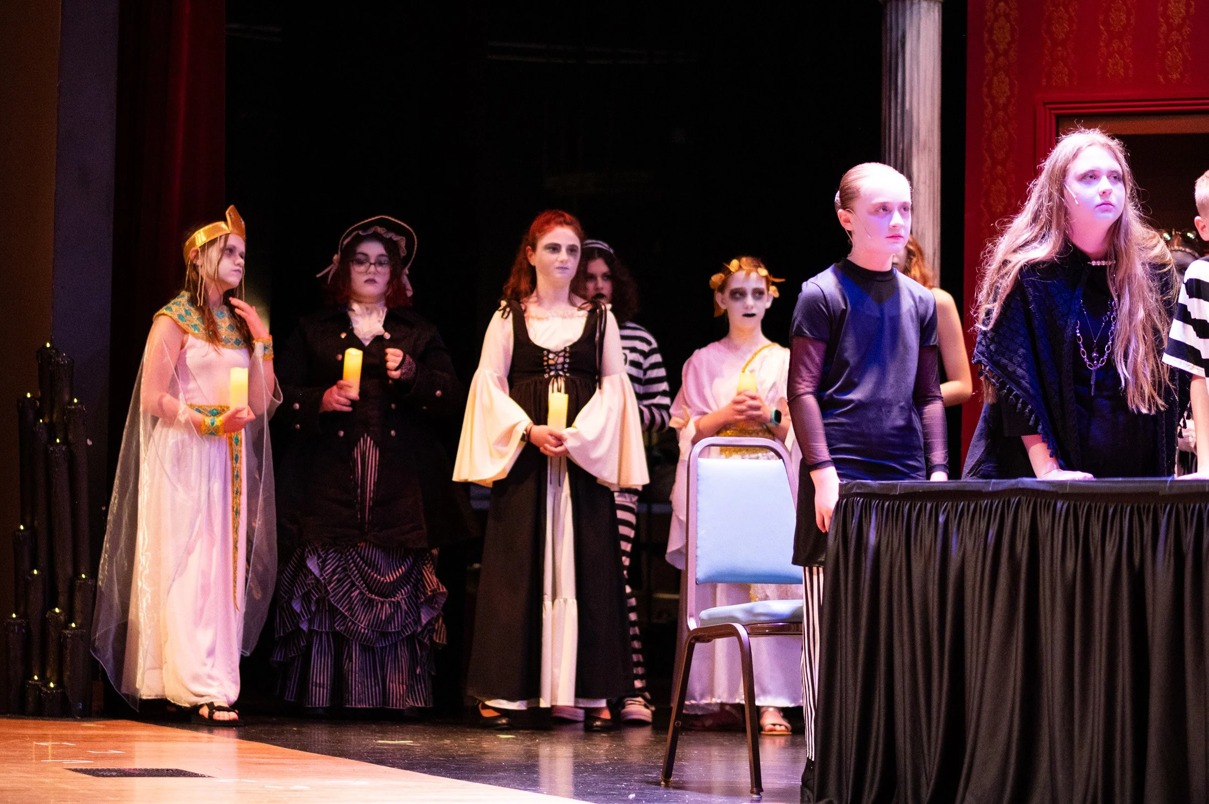 Children in costumes stand on stage, holding candles, during a theatrical performance with a dark backdrop and a red curtain on the side.